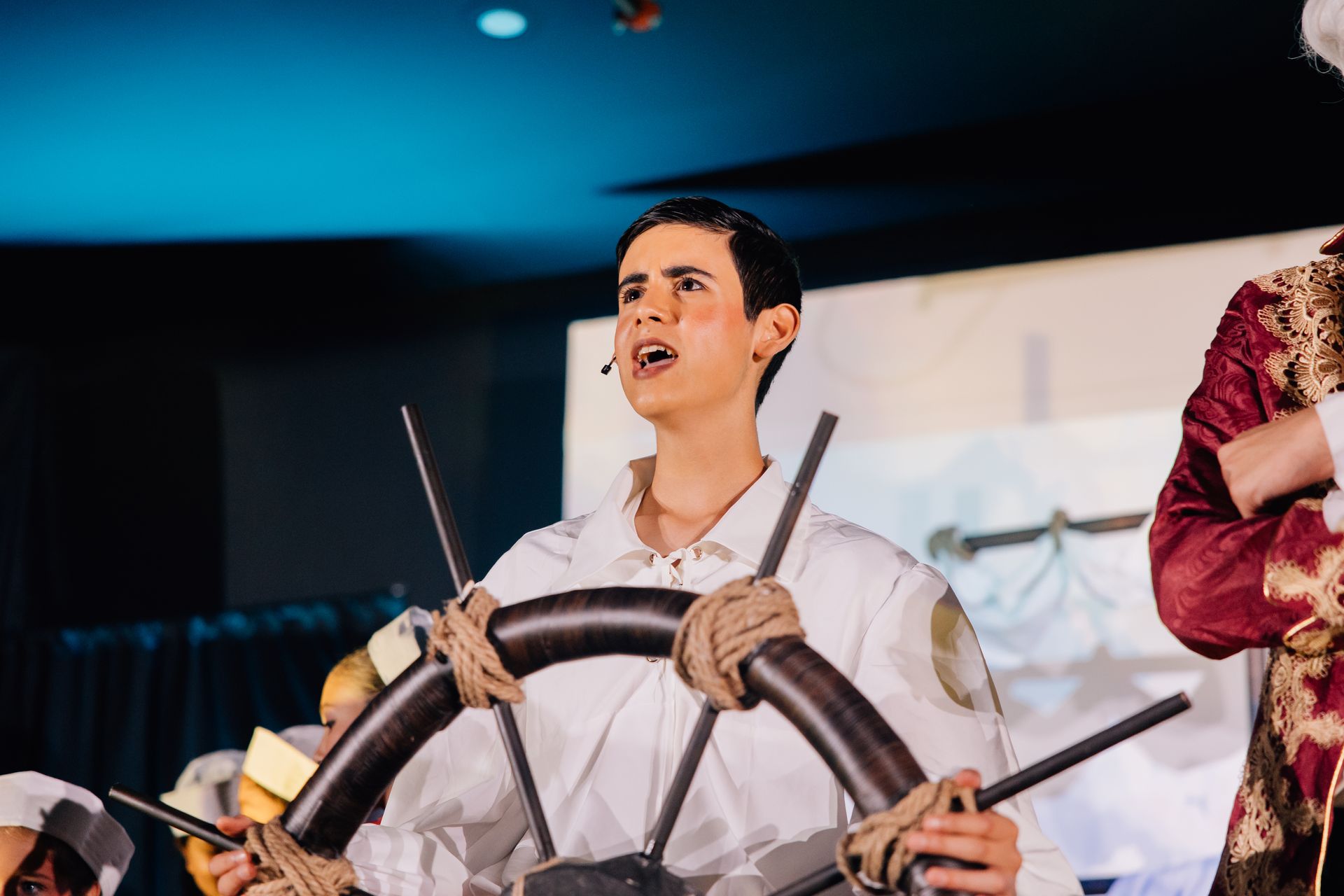 Man in white shirt holding a ship’s wheel on stage under blue lighting