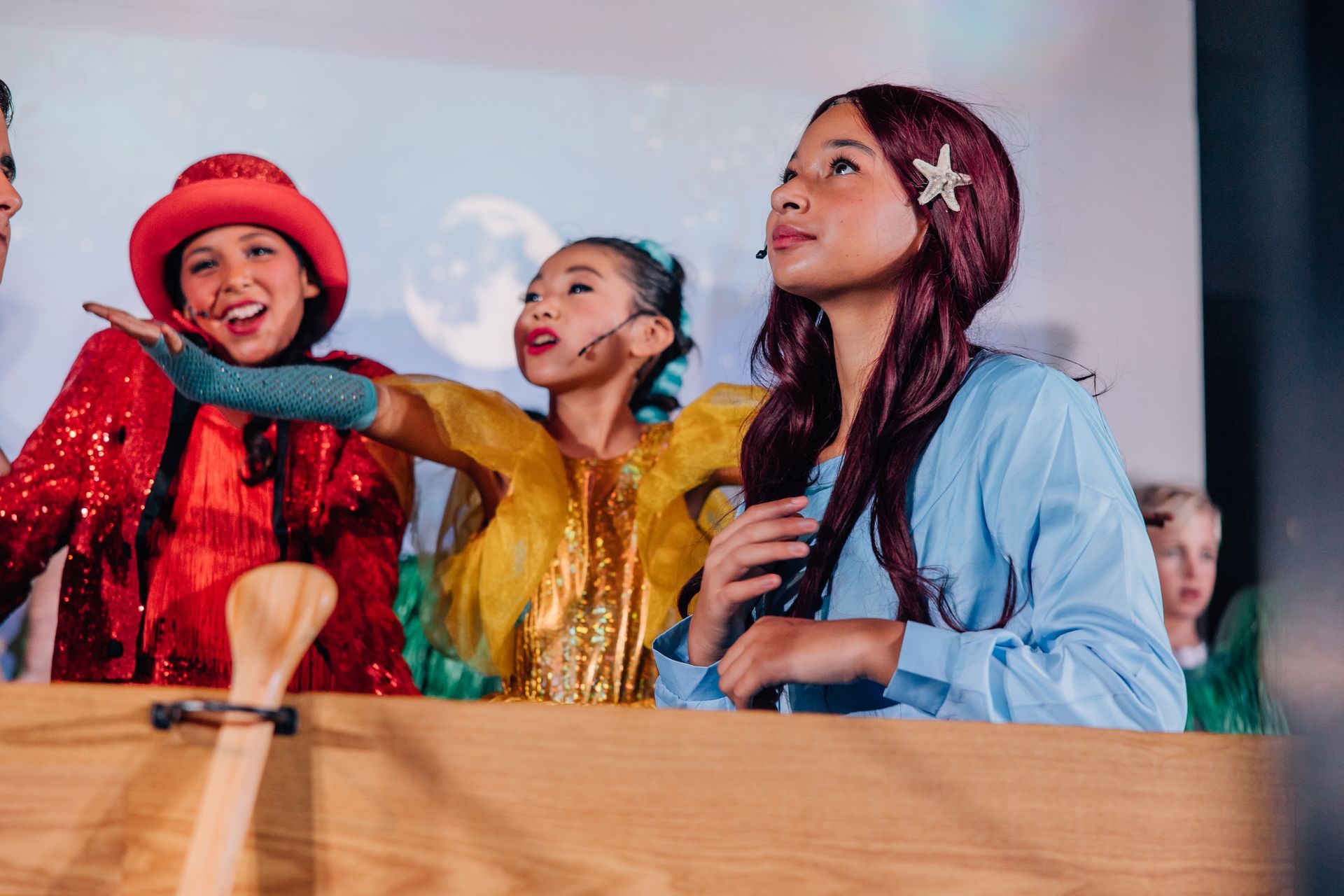 Three performers in colorful costumes singing on stage behind a wooden barrier for the  Little Mermaid stage production.