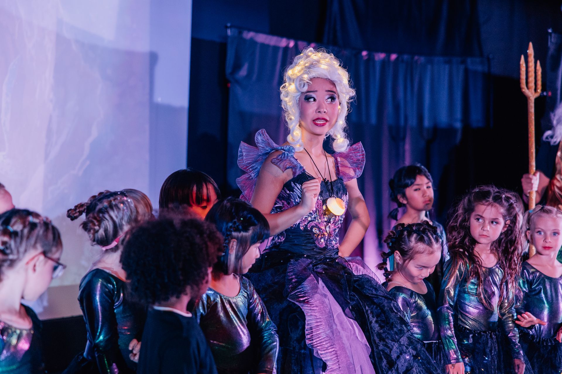 Costumed performer onstage with children, wearing a black and purple outfit  for the  Little Mermaid stage production.