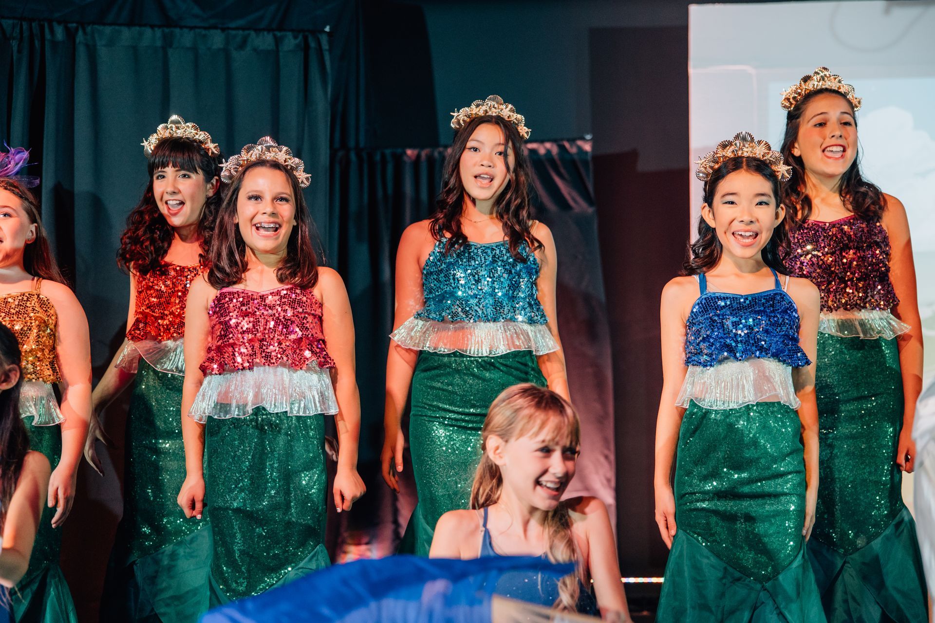 Children in sparkly mermaid costumes performing on stage with curtains behind them for the  Little Mermaid stage production.