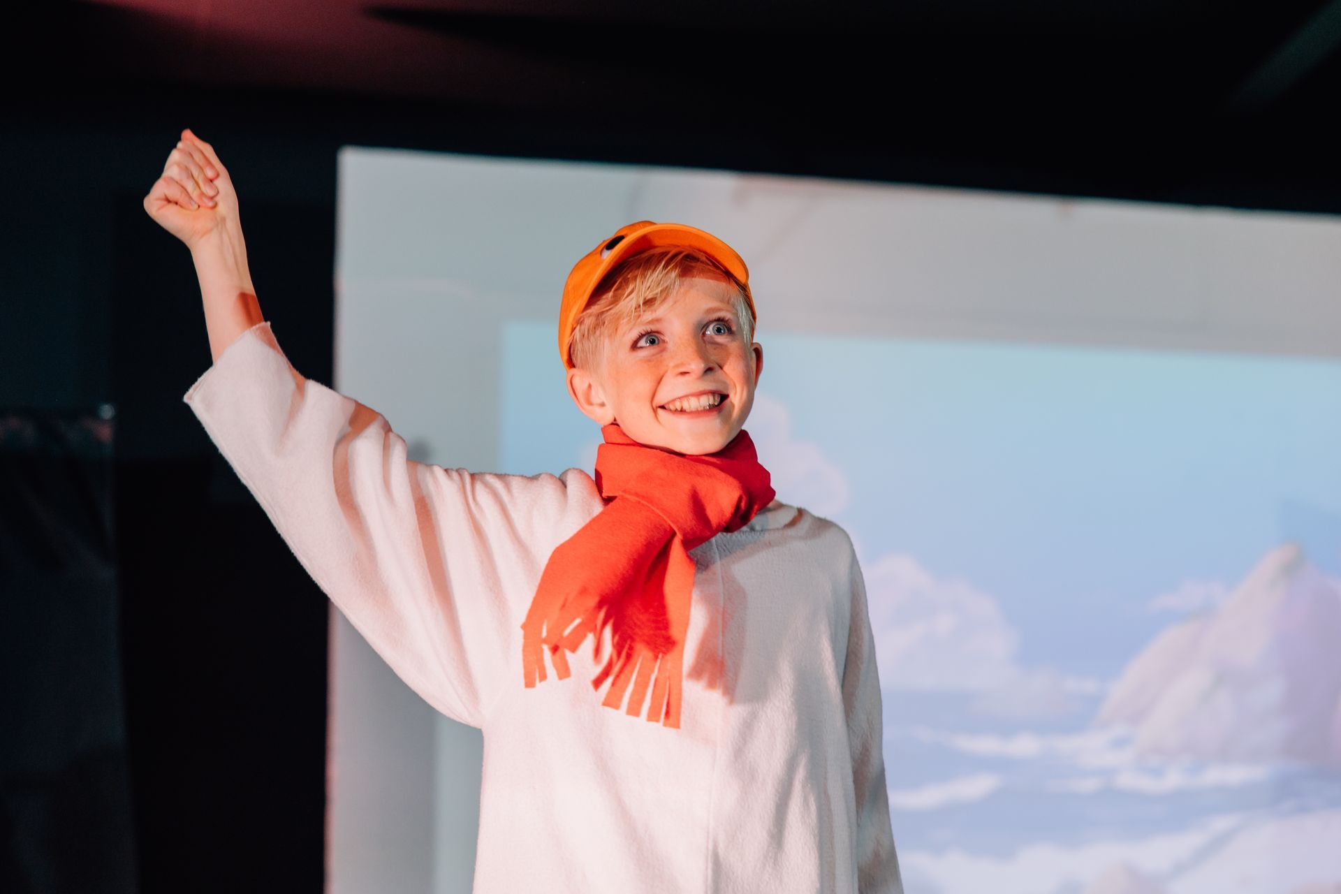 Smiling performer in orange hat and scarf raises one arm onstage before a projected snowy backdrop