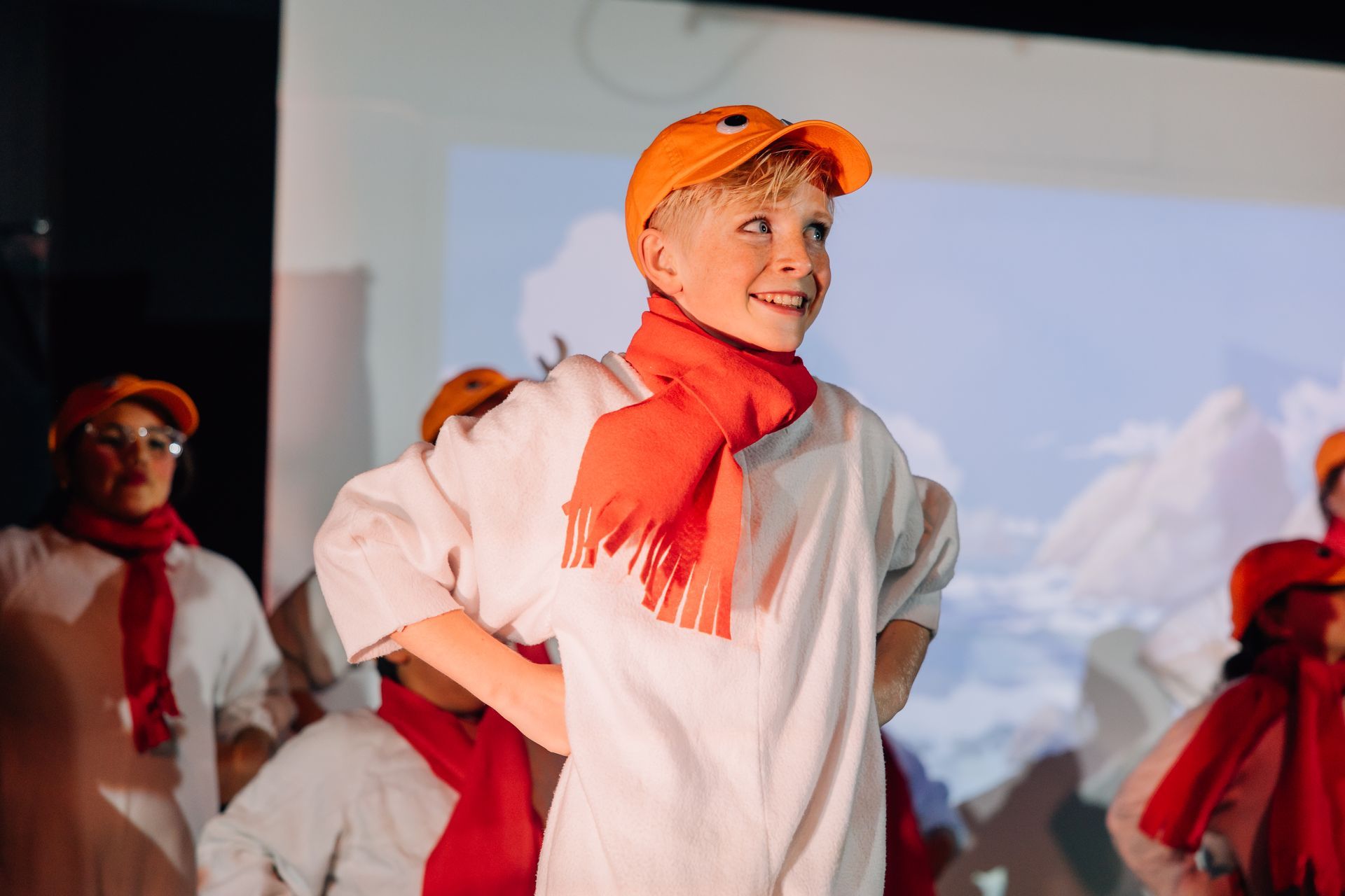 Children in red scarves and hats perform on stage, smiling under warm lights for the Little Mermaid stage production.