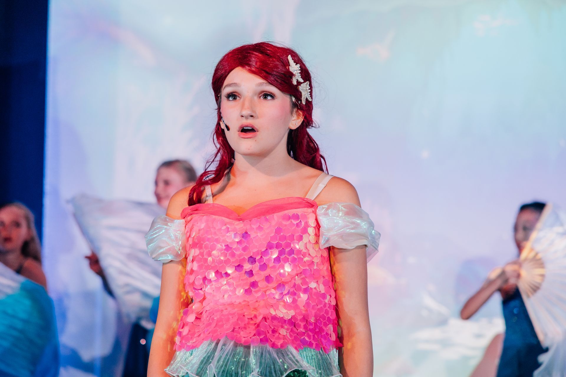 Child in a pink sequined dress with red hair stands onstage for the  Little Mermaid stage production.