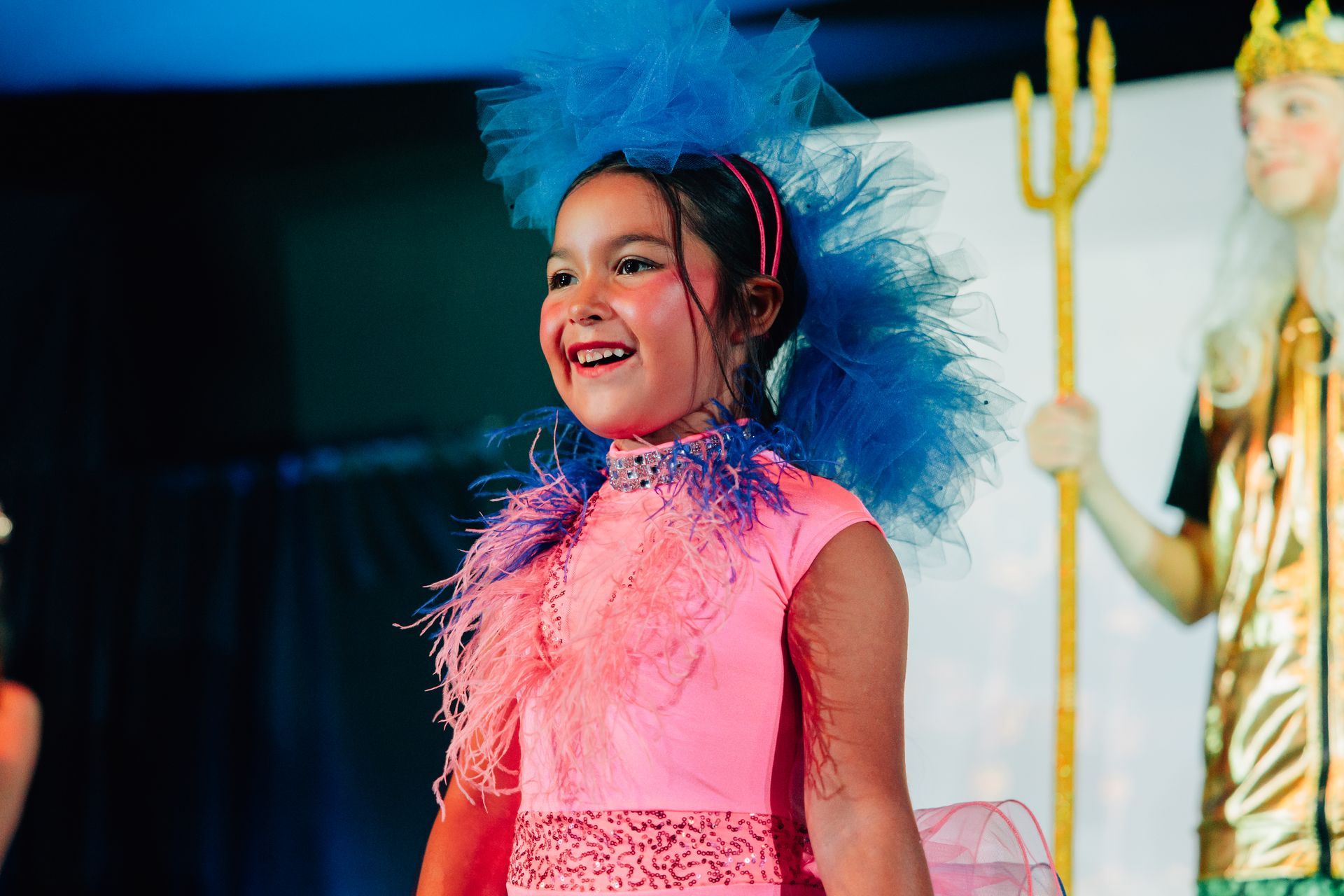 Smiling child in a pink costume with blue feathers on stage for the  Little Mermaid stage production.