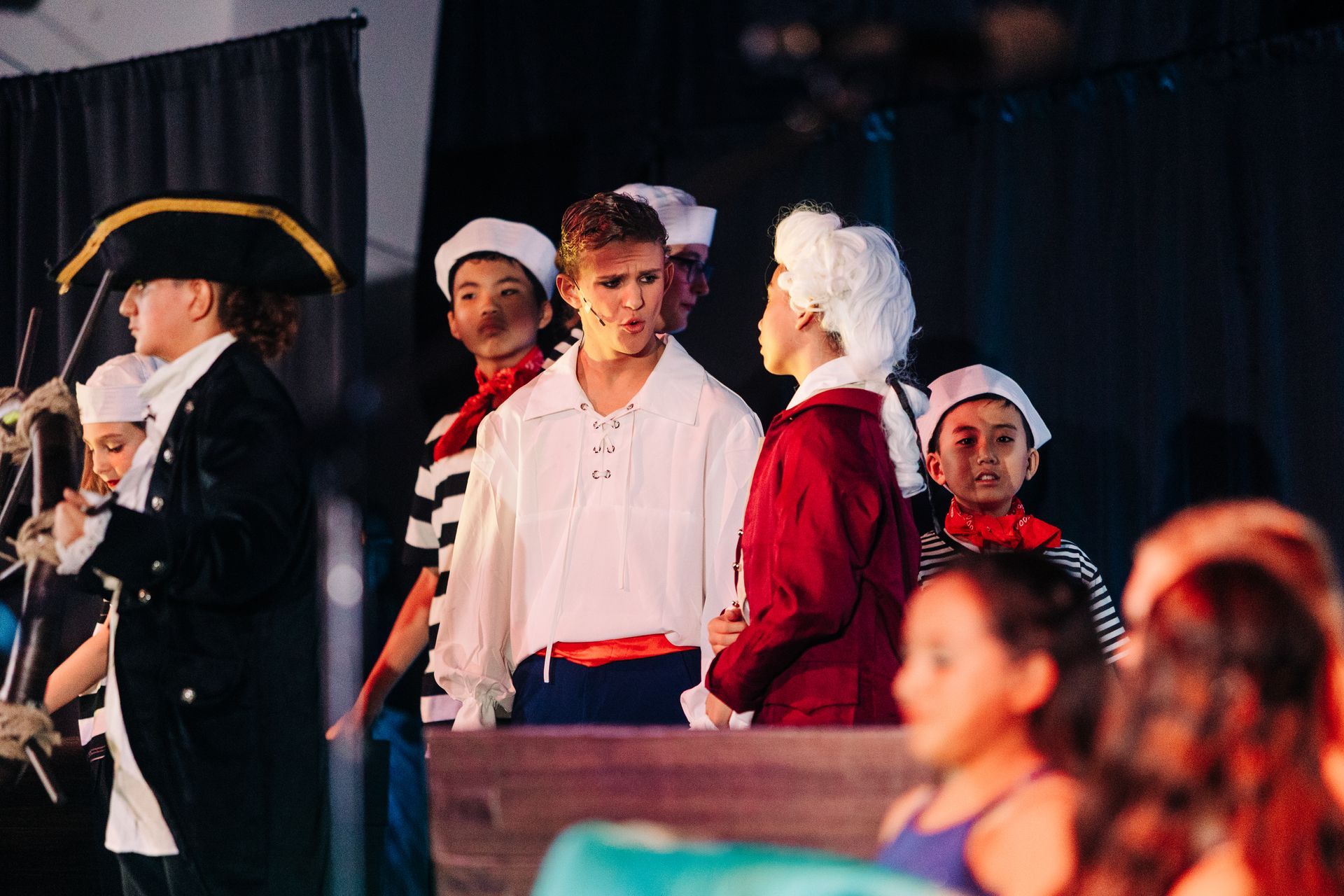 Actors in period costumes on a theater stage, with a man in a white shirt centered under warm lights.