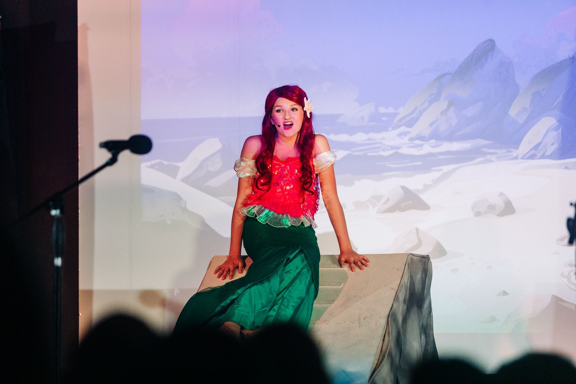 Woman in red top and green skirt posing on stage before a snowy mountain backdrop