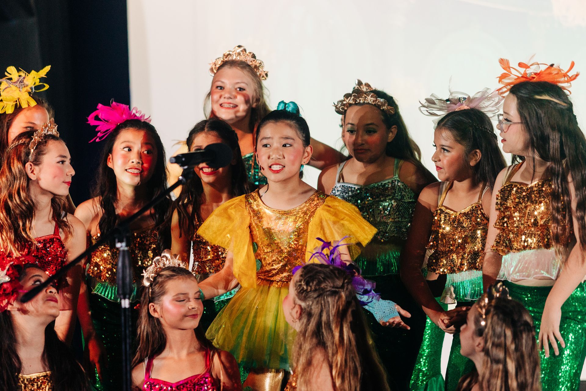 Children in colorful costumes and headpieces performing on stage, smiling and singing together