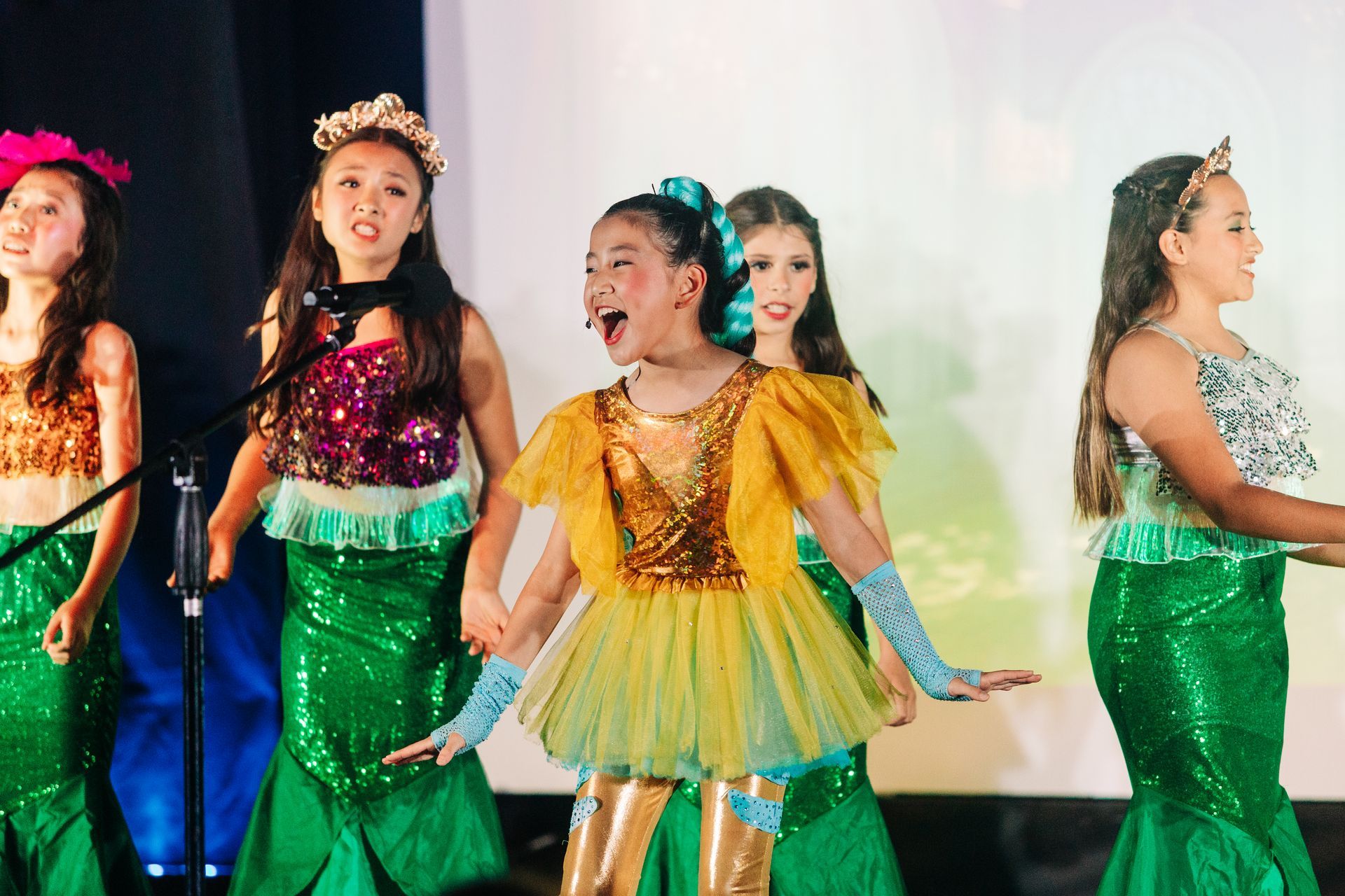 Performers in glittery green and yellow costumes singing onstage under bright lights