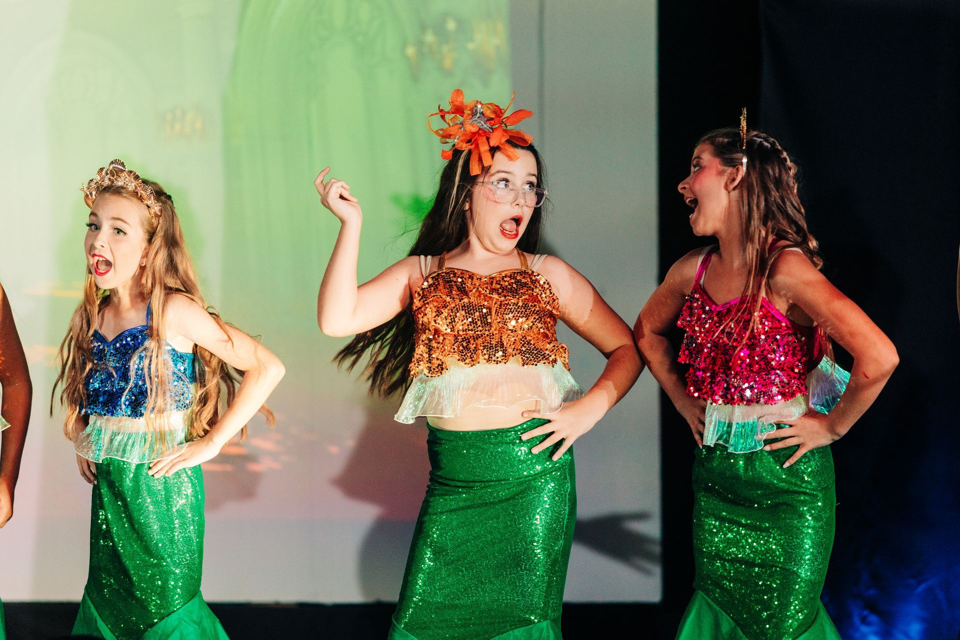 Three dancers in green skirts and sparkly tops performing onstage with hands on hips