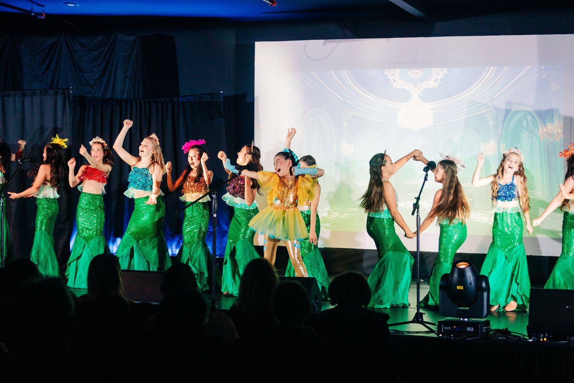 Dancers in green costumes perform on a stage with colorful lights and a projected backdrop.