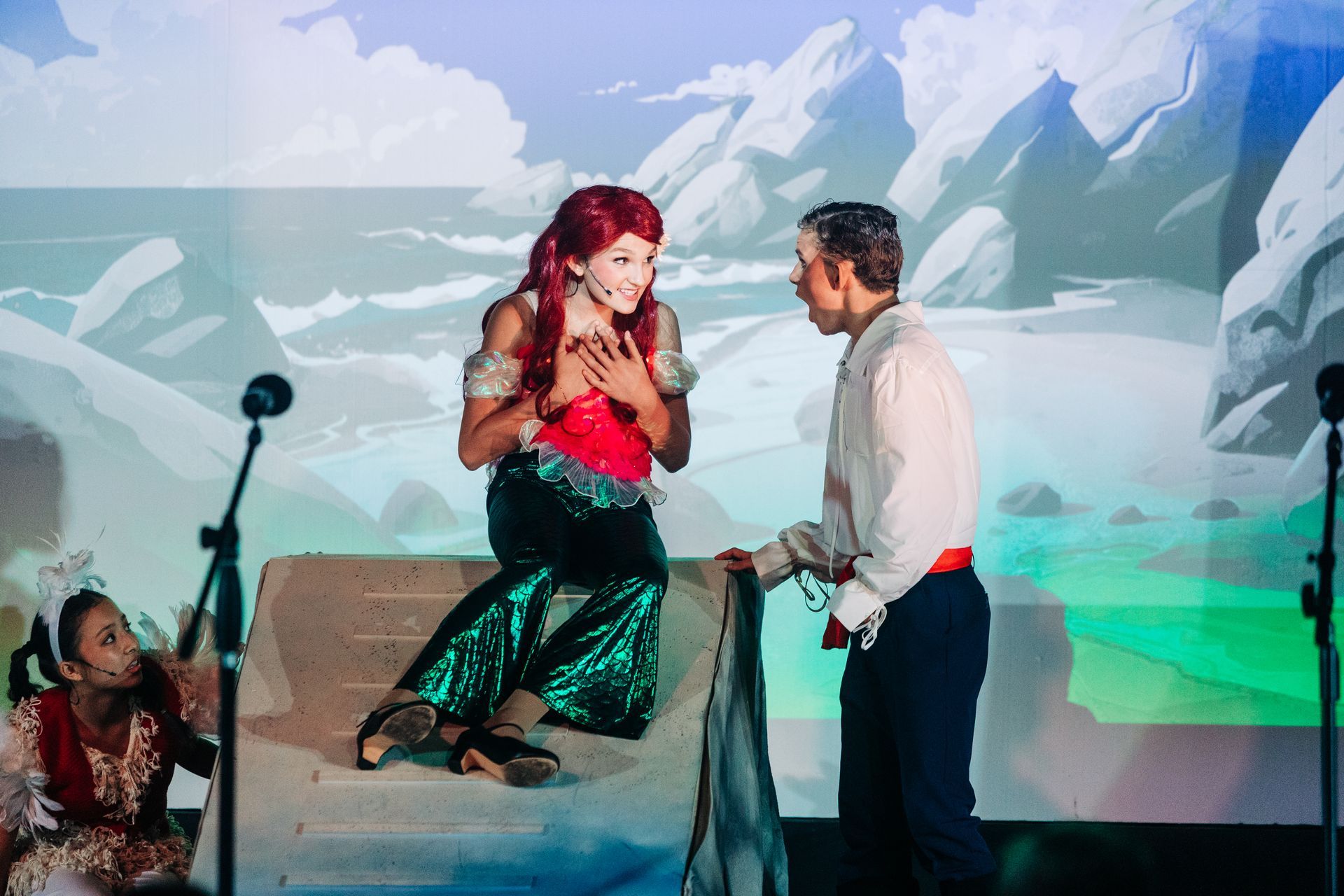 Actor in red and green costume performs on stage beside another actor, with painted mountain backdrop.