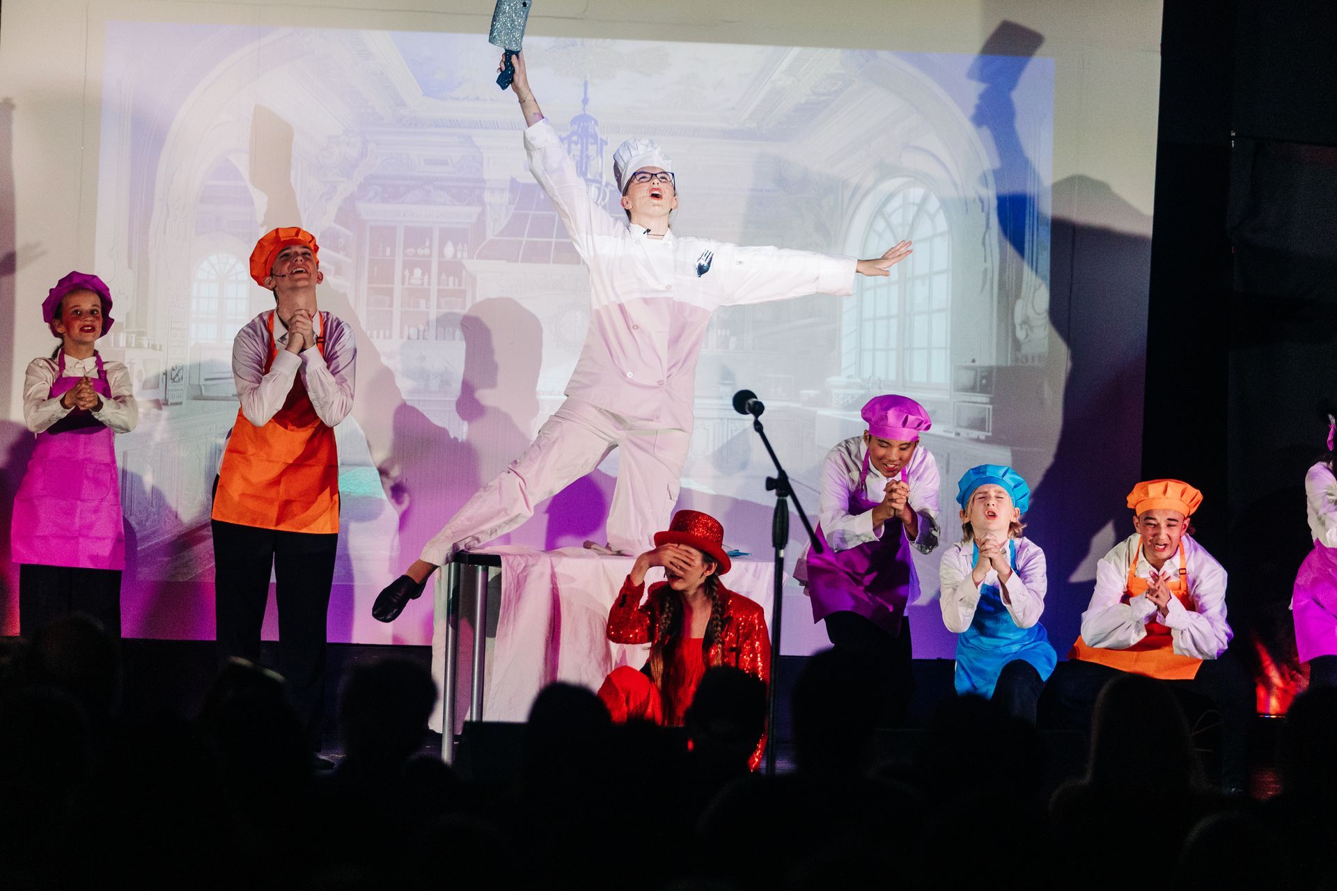 Stage performance with colorful-costumed performers, one in white leaping center under a projected backdrop.