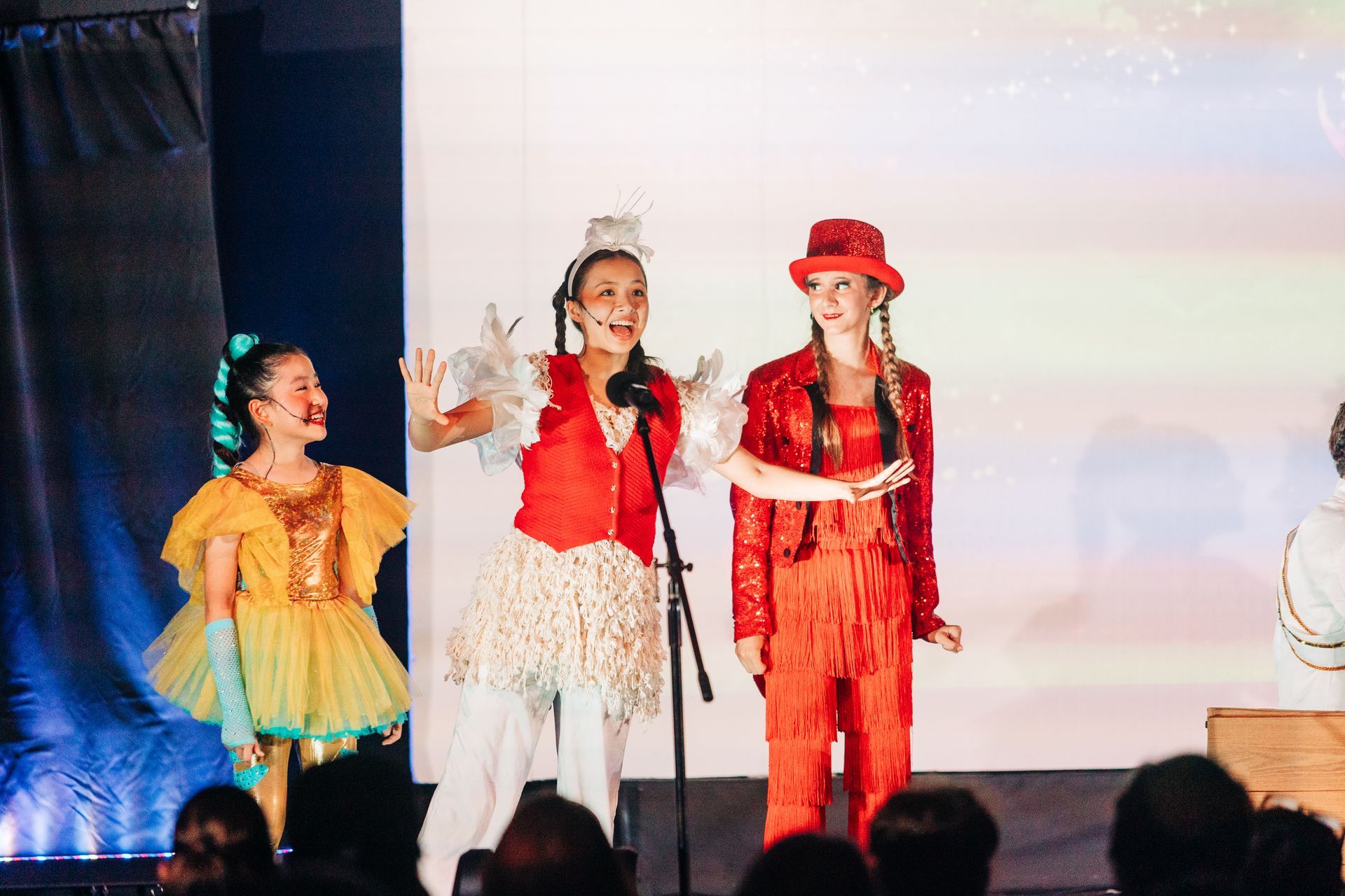 Three costumed performers on stage, two singing into microphones under bright lights.