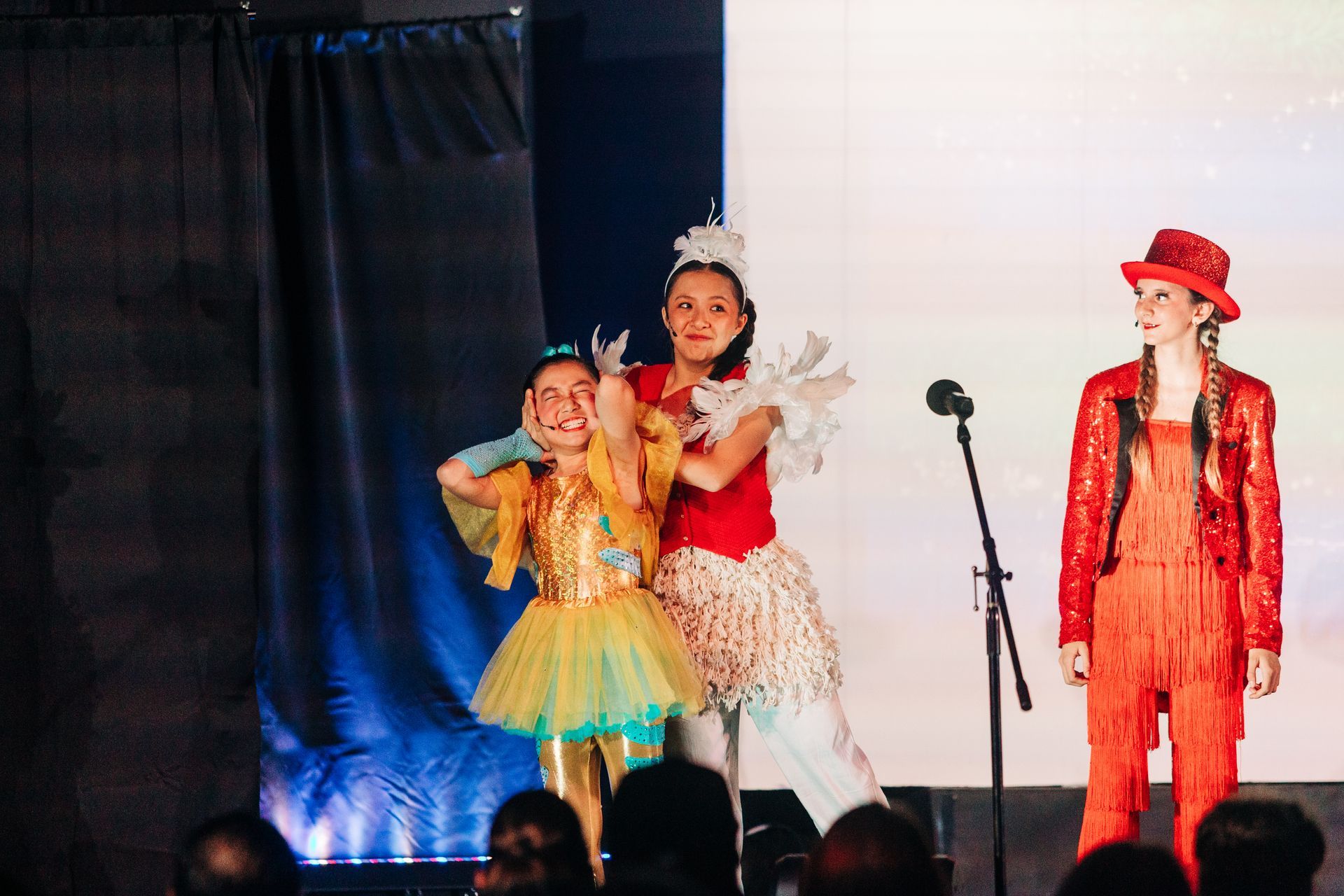 Three children in colorful costumes posing on a stage with a microphone stand and curtain backdrop