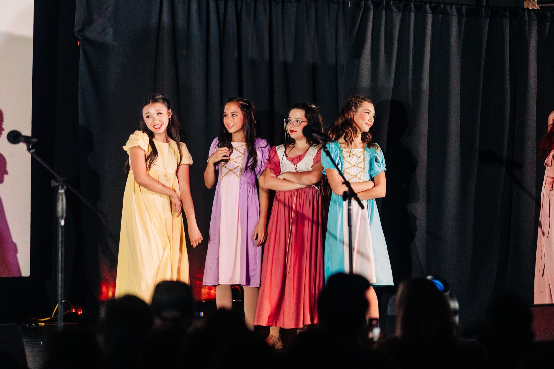 Four performers in colorful dresses stand on stage singing into microphones before a black curtain.