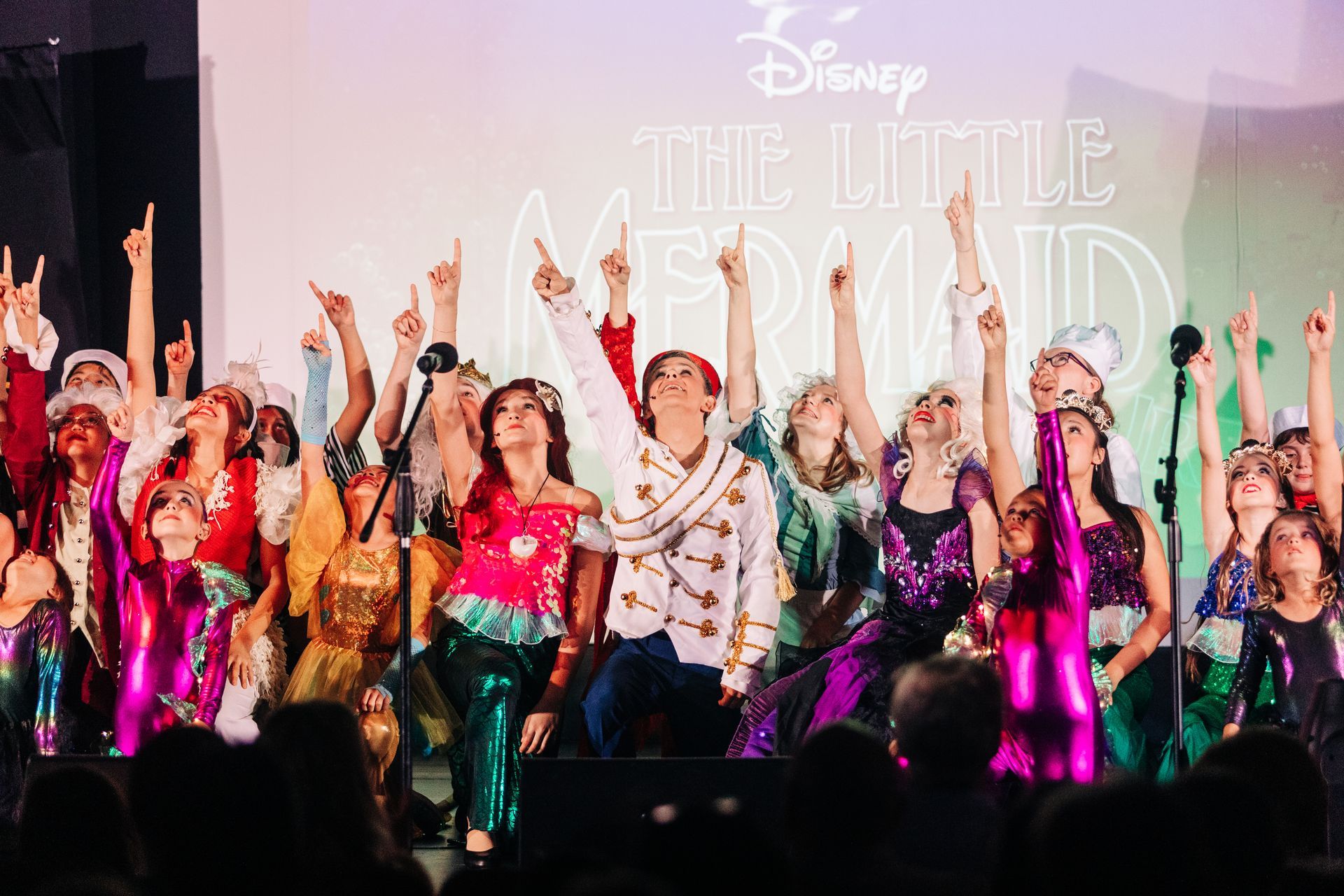 Disney cast in colorful costumes dancing on stage in front of a Little Mermaid backdrop
