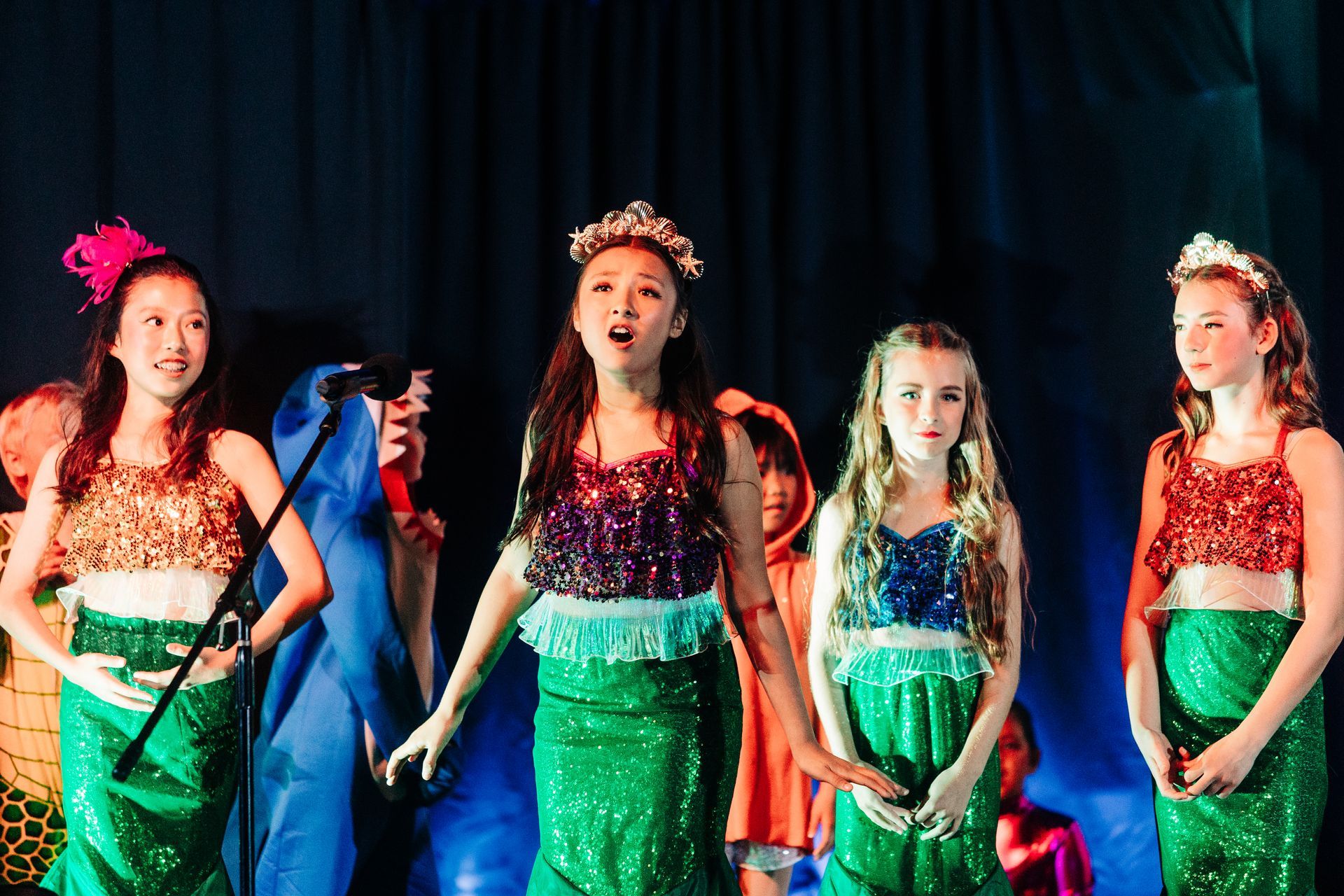 Four performers in sparkly green costumes stand onstage under colorful lights