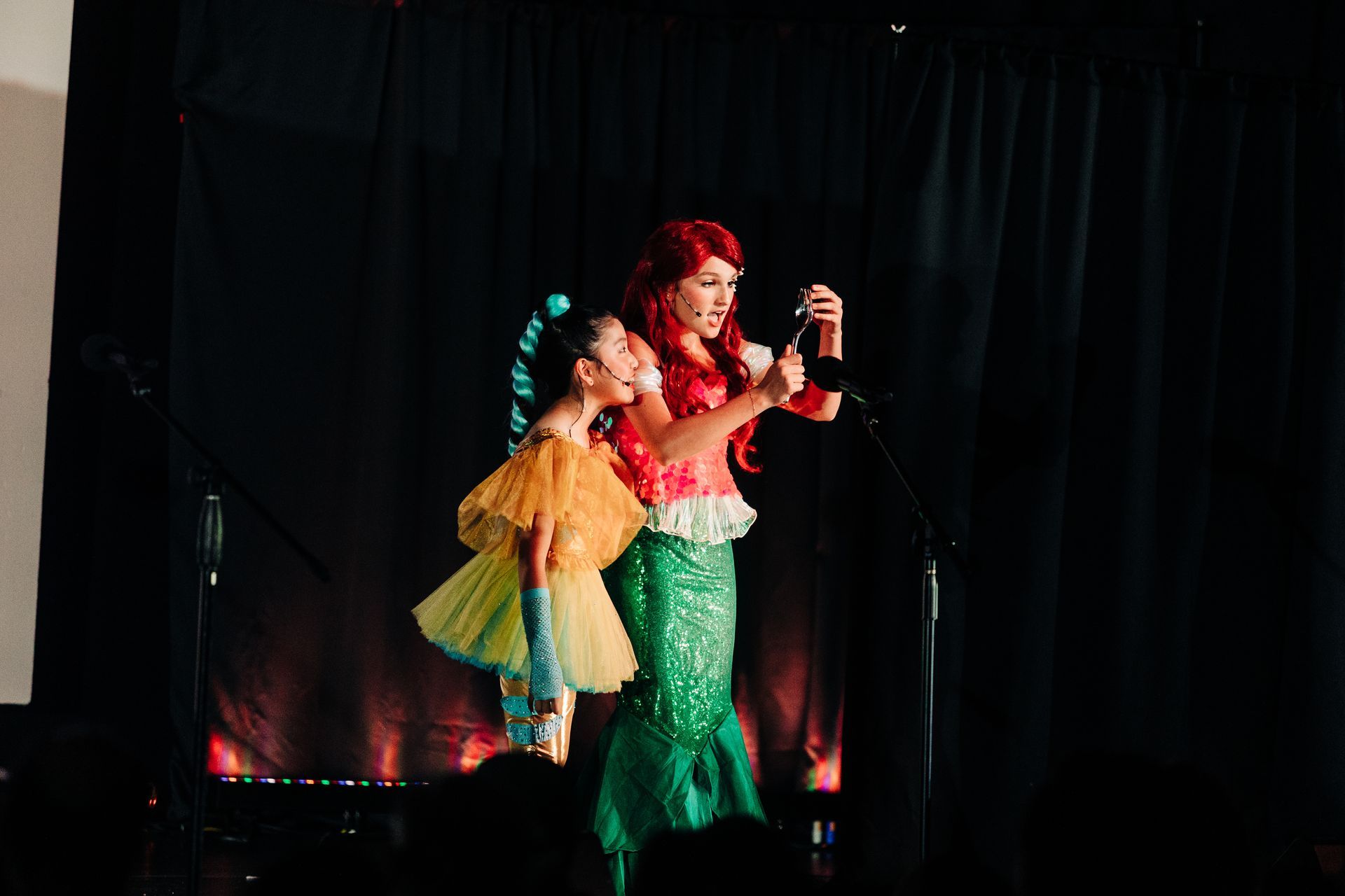 Two performers onstage in colorful costumes, singing under warm lights against a black curtain