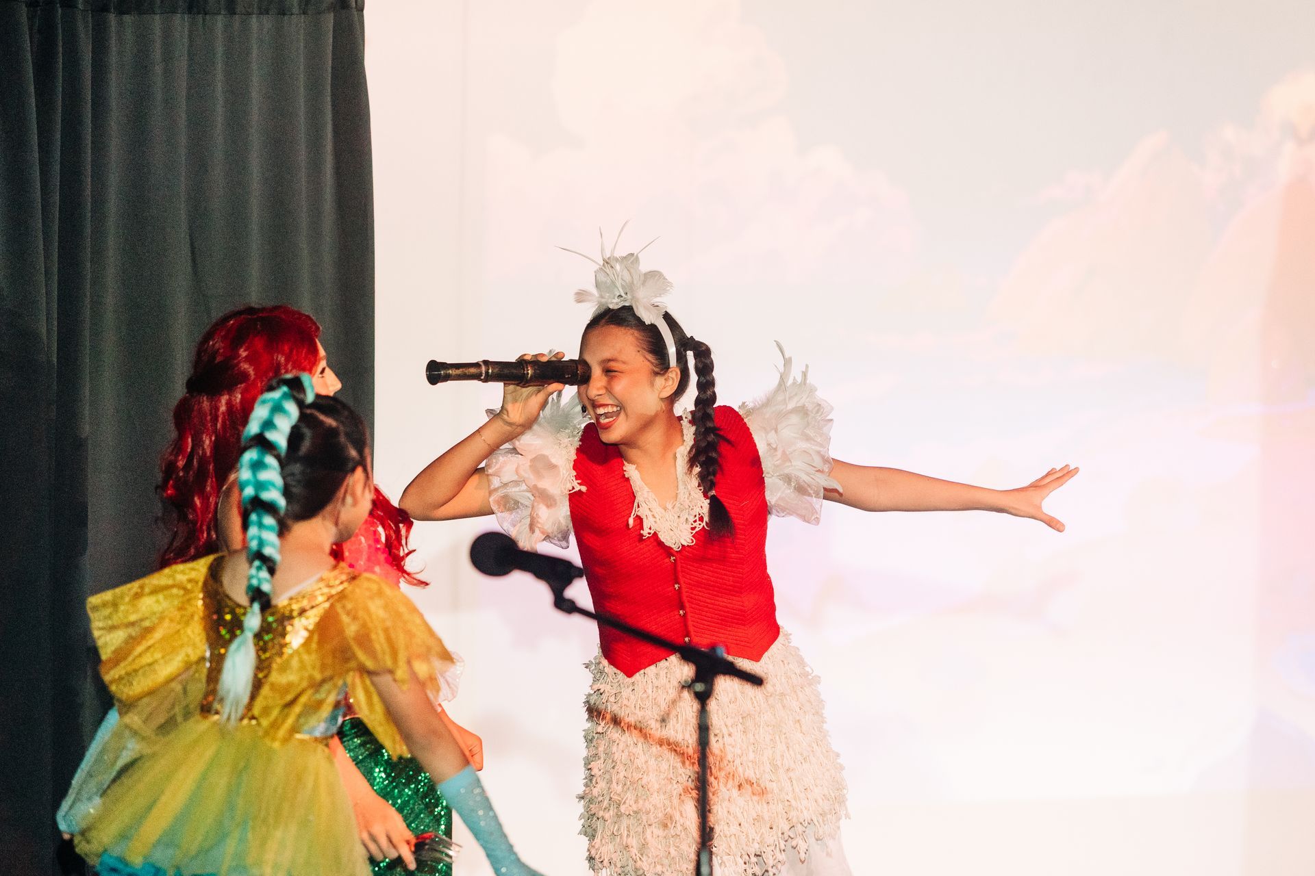Two children in colorful costumes performing onstage, one singing into a microphone while the other reaches out.