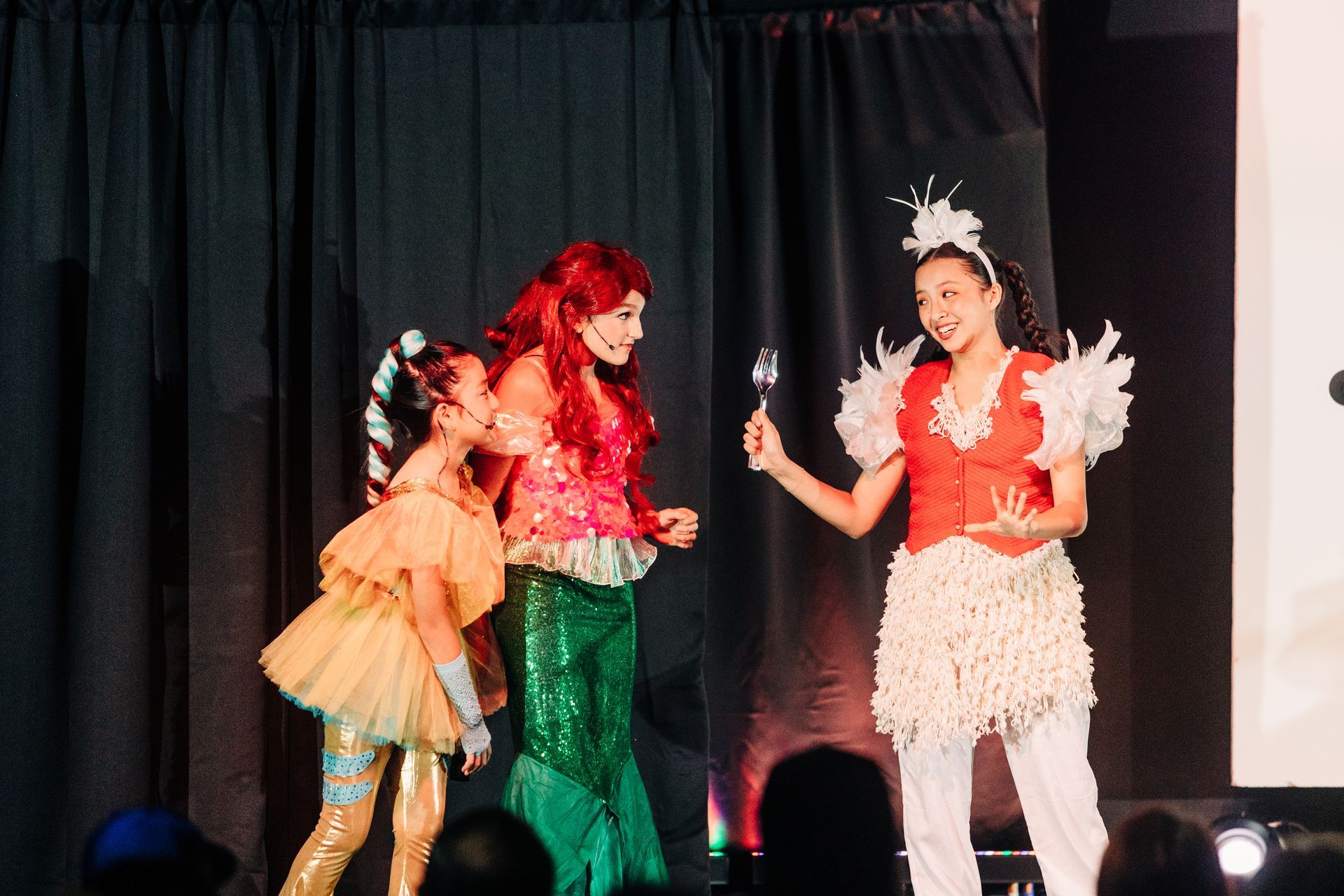 Three costumed performers posing on a stage, with colorful outfits and a black curtain backdrop
