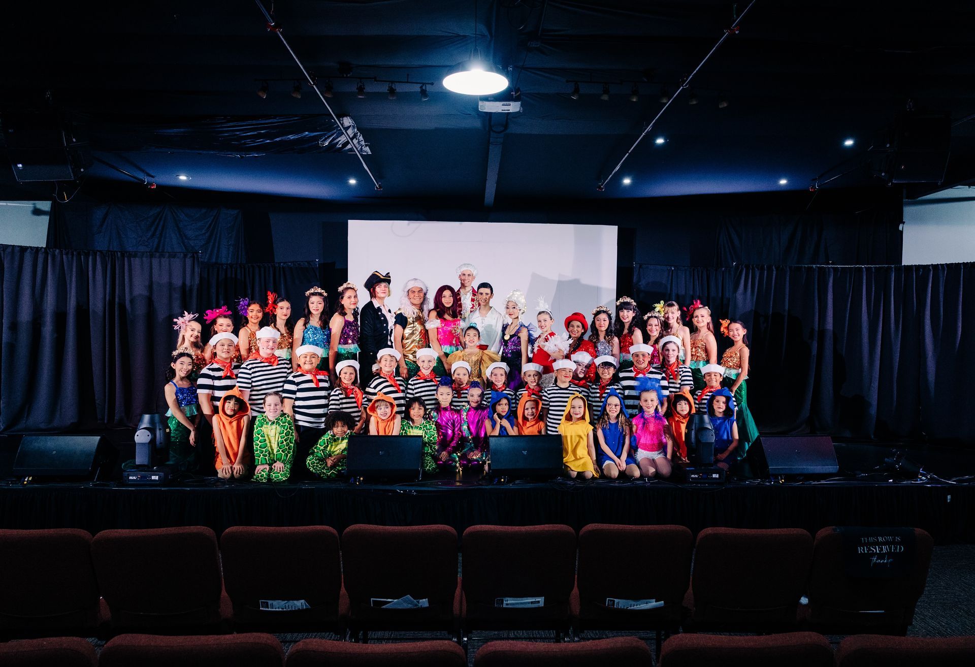 Large group posing on a theater stage with colorful costumes and a blank screen behind them