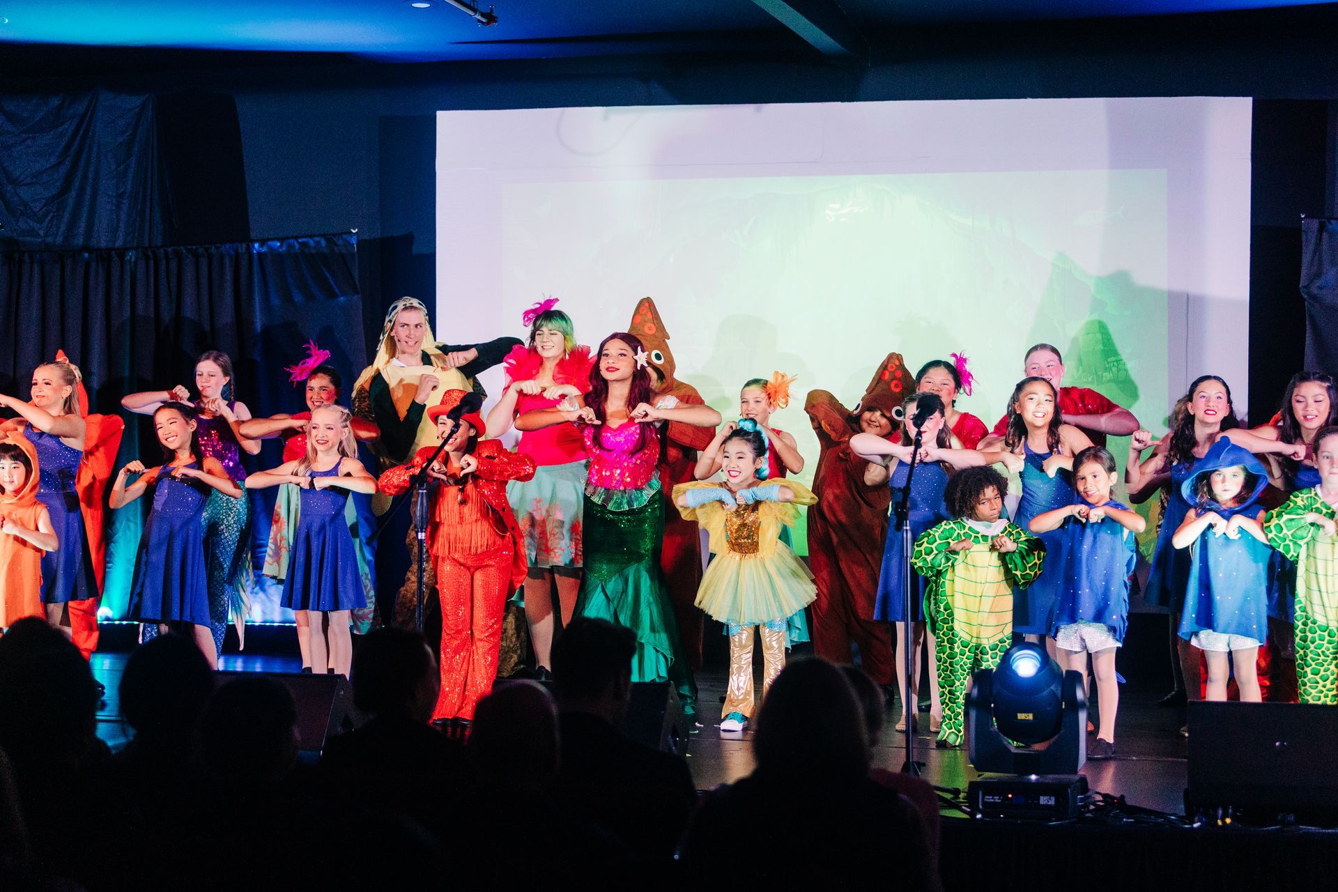 Children in colorful costumes performing on a stage with bright lights and a projection screen behind them