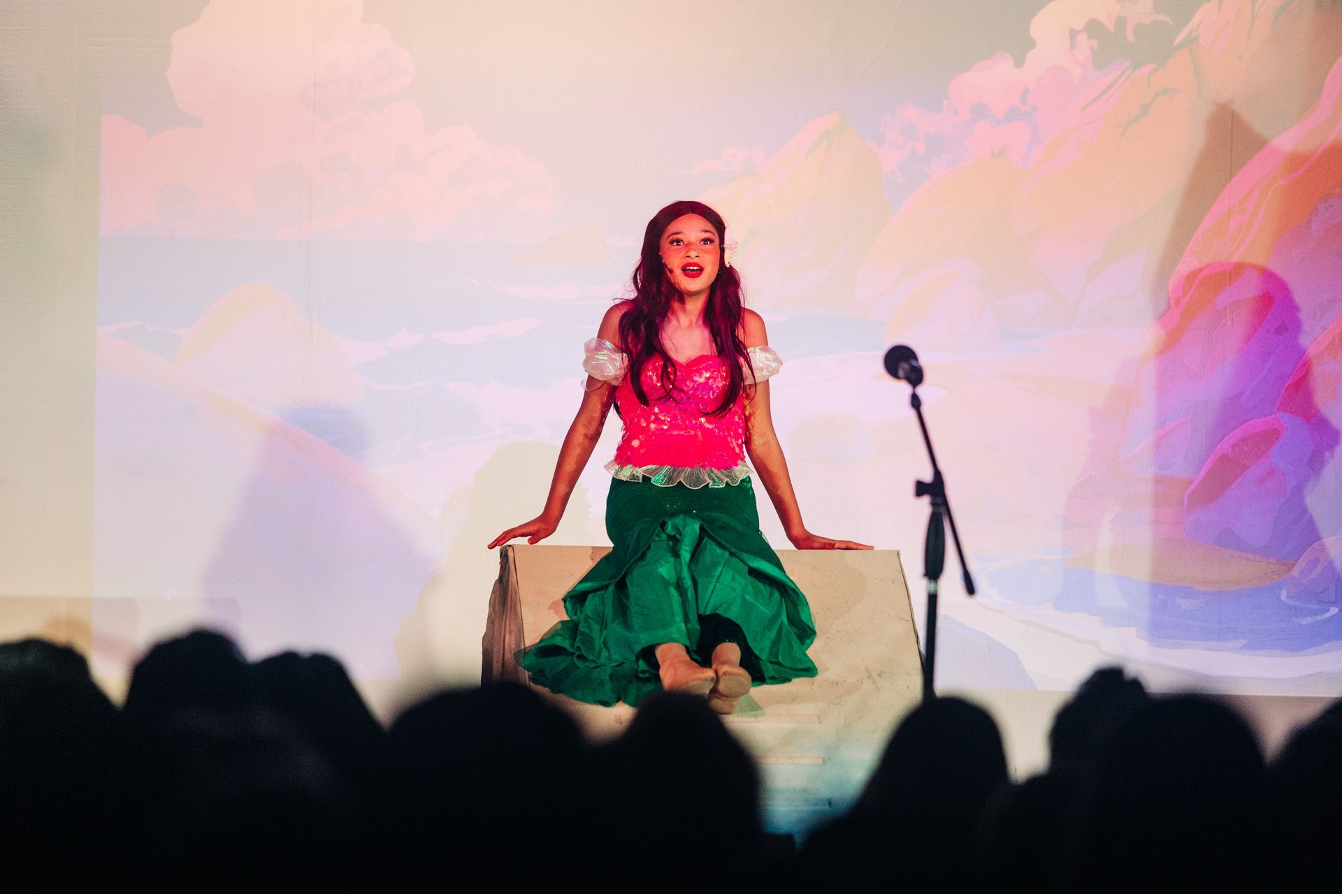 Woman in a pink top and green skirt sits on a stage, with a microphone stand beside her and audience silhouettes below.
