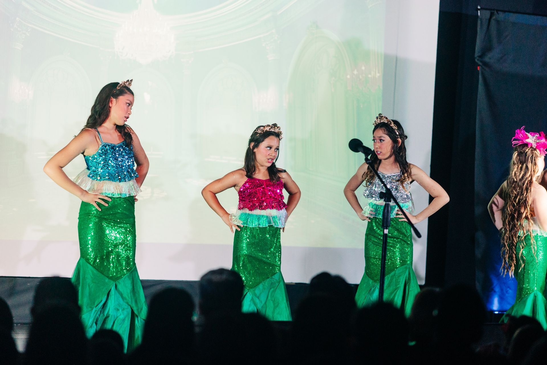 Four mermaid-costumed performers on stage, posing under stage lights with an audience silhouette in front.