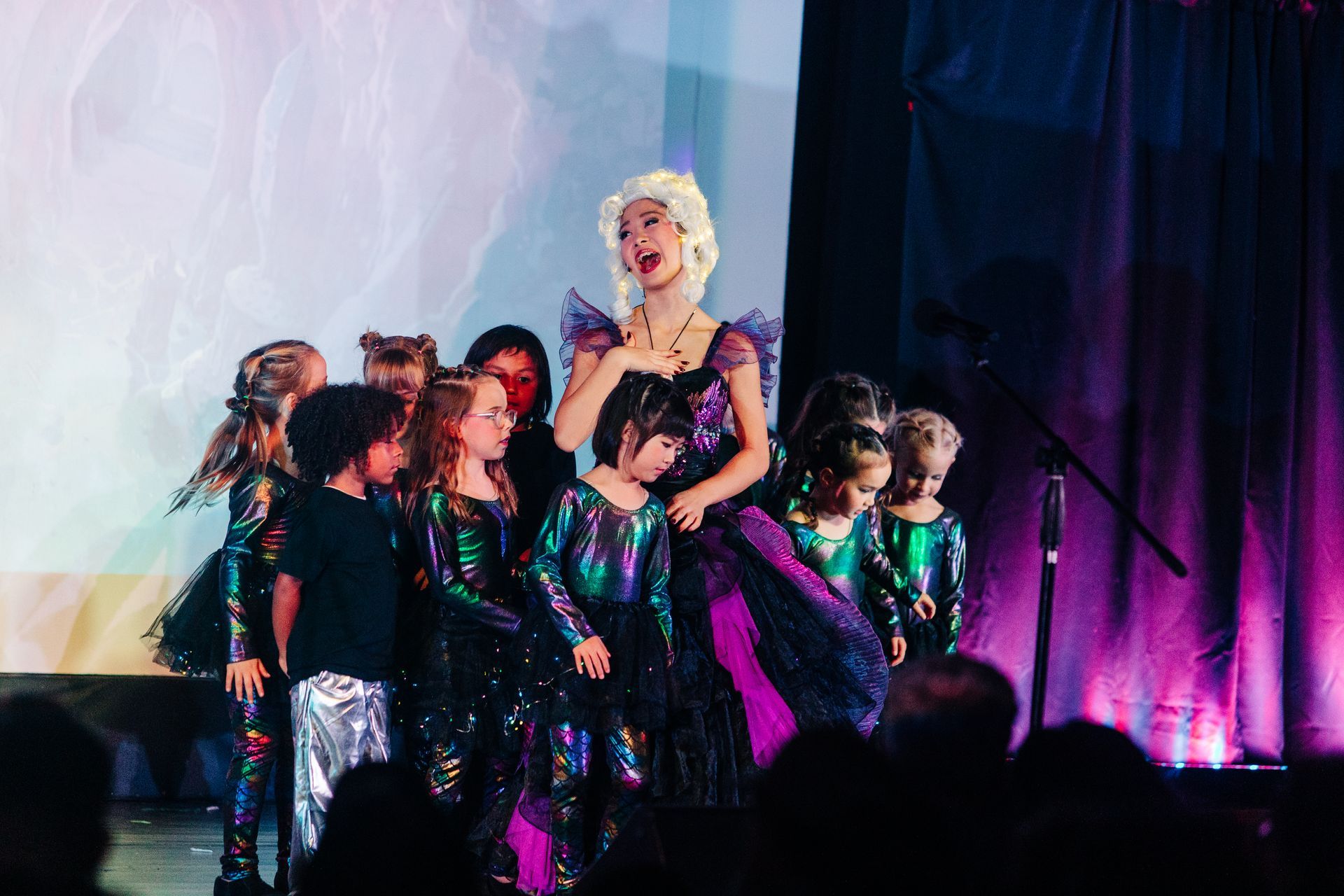 Performer in a glittering dress poses center stage with dancers in black outfits under purple lighting