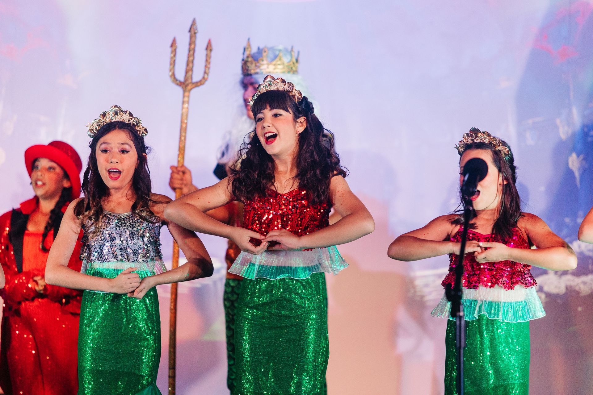 Three performers in green skirts and festive headpieces sing onstage; one holds a trident.