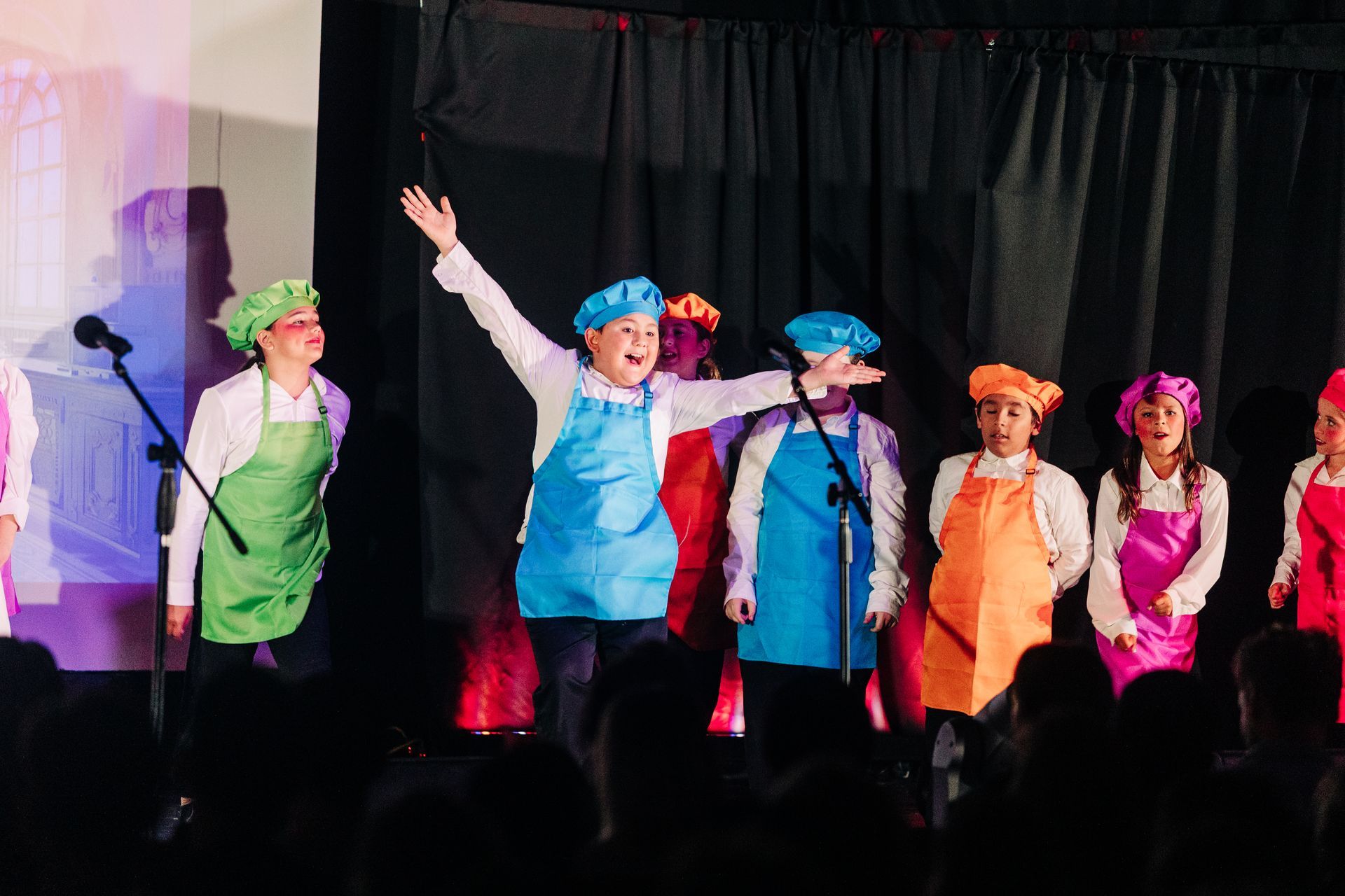 Colorful costumed performers dancing on stage with arms raised under black curtains