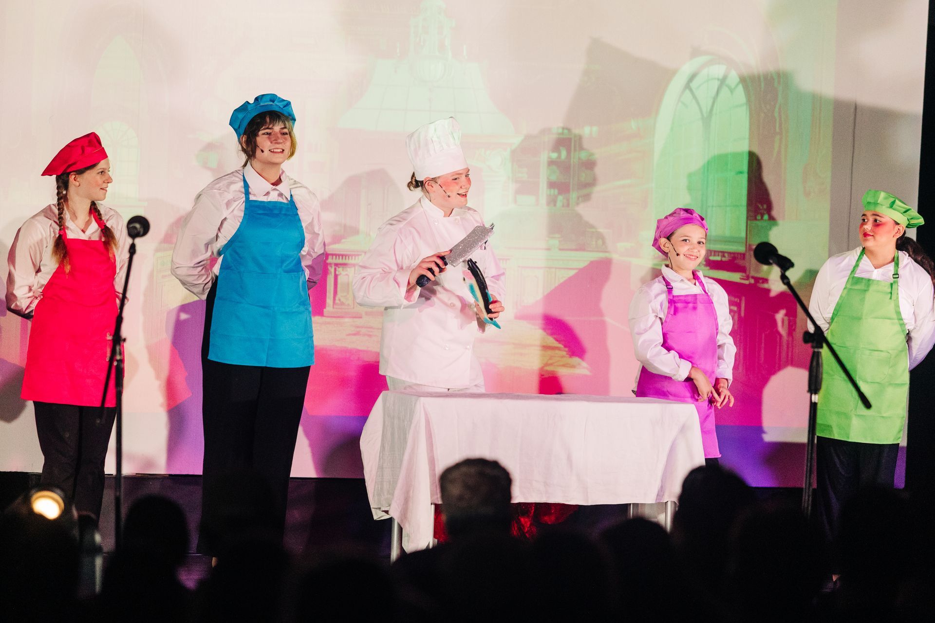 Five performers in colorful costumes on a stage, standing around a table under pink and green lights
