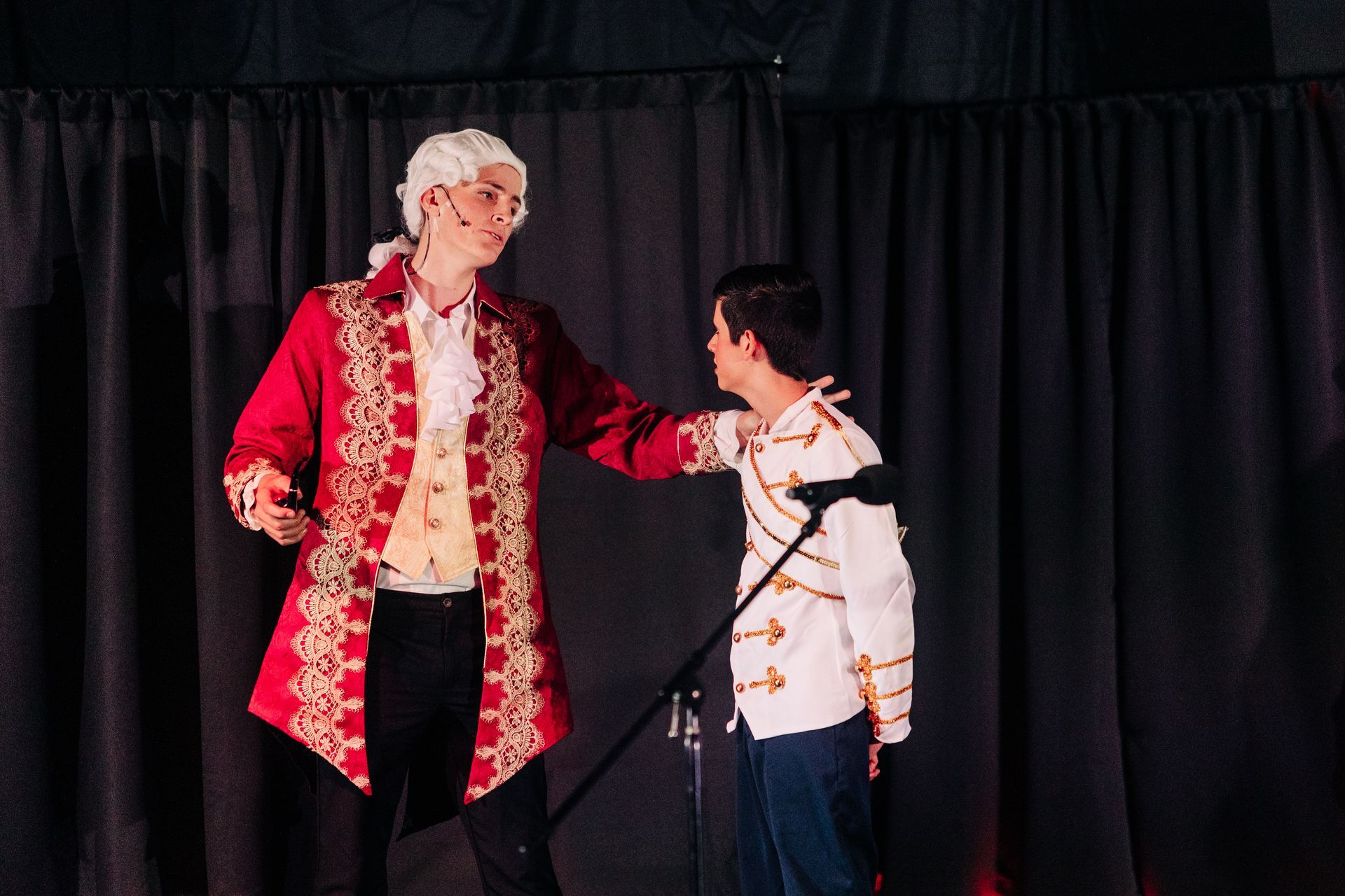 Two performers onstage in costume, one in a red coat speaking to another by a microphone.