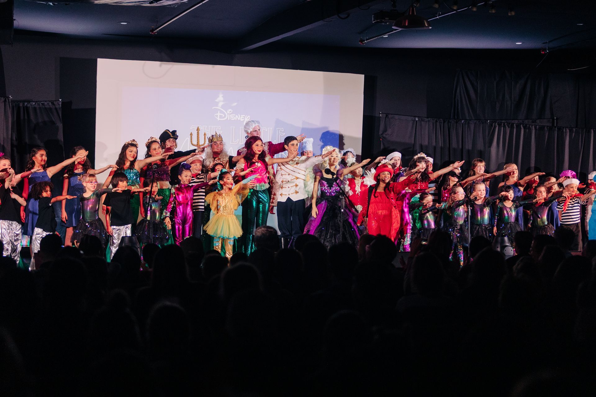 Group of dancers performing onstage in colorful costumes with arms raised under stage lights