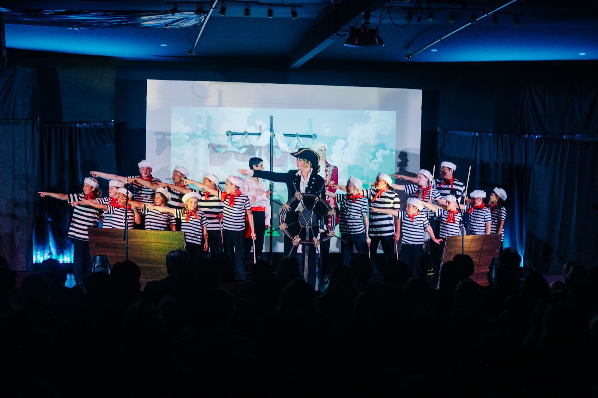 Children in striped shirts perform on stage with a conductor under blue lighting.