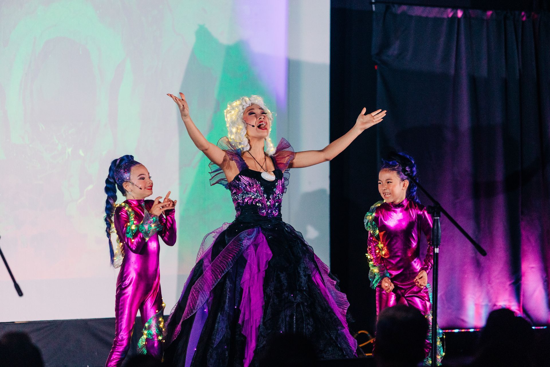 Three performers in glittering purple costumes on a lit stage, center performer with arms raised