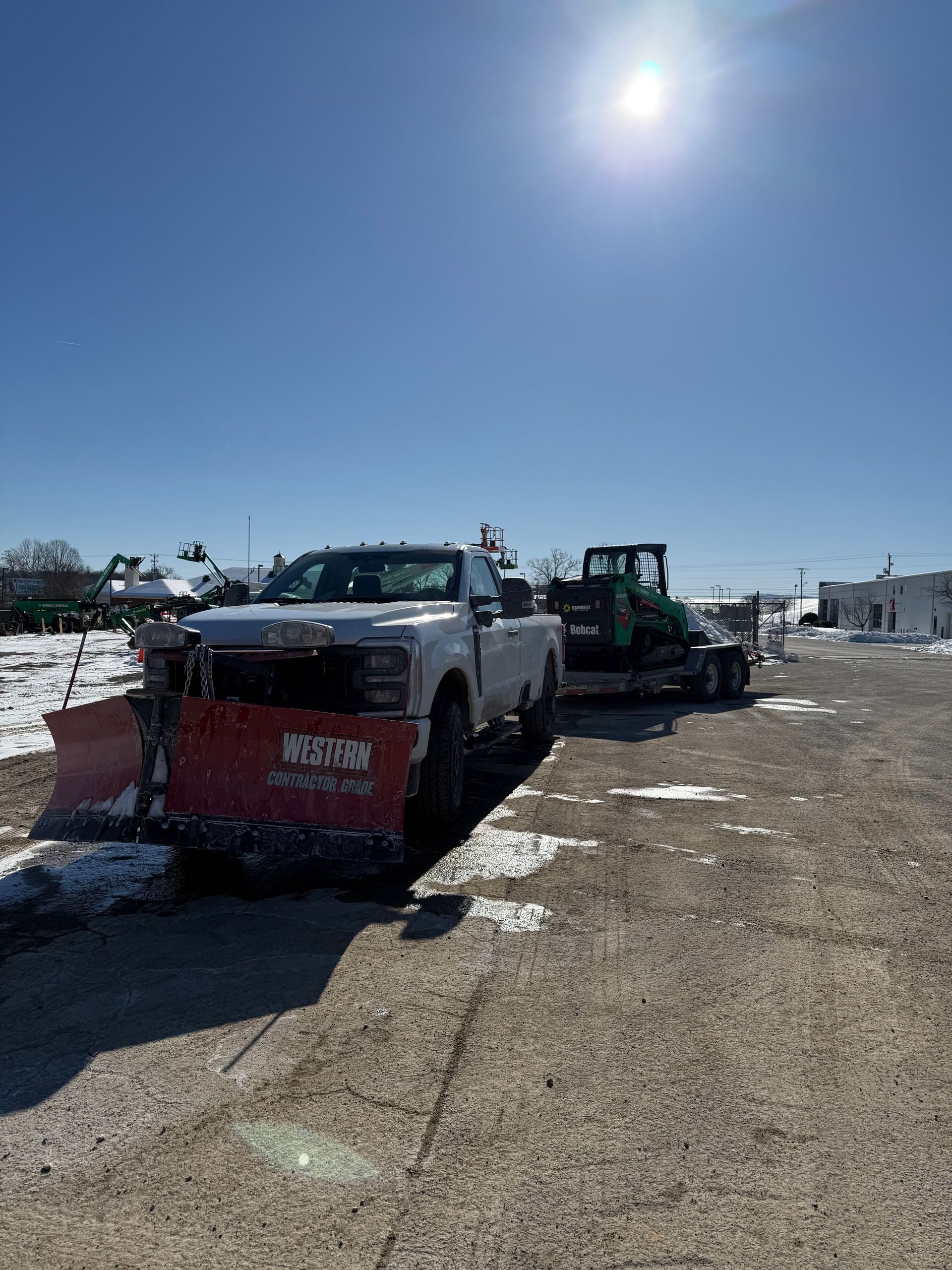 Snowplow truck and skid steer on a muddy, snow-covered road under a bright sun