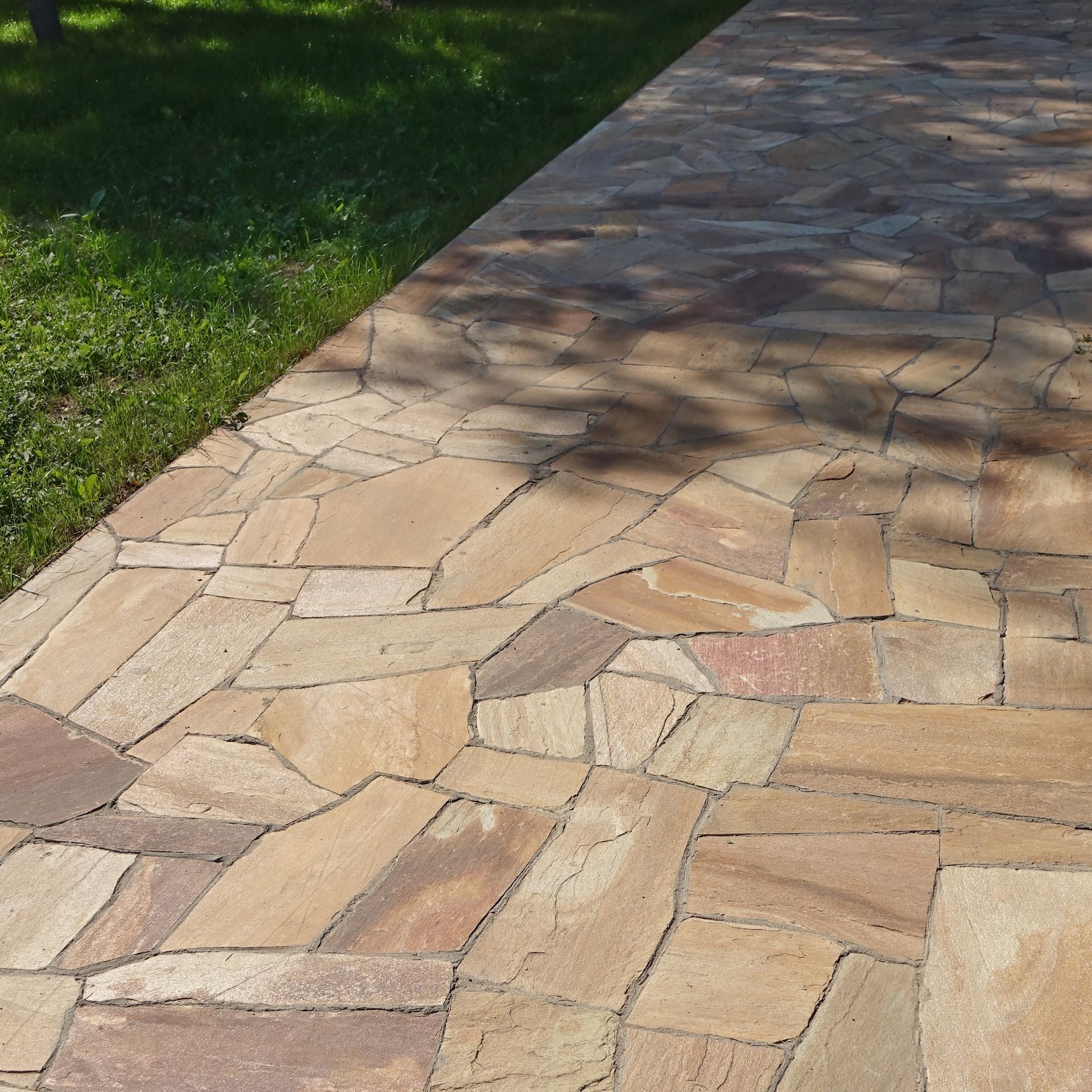 Stone walkway beside green grass in sunlight, with tree shadows across the path