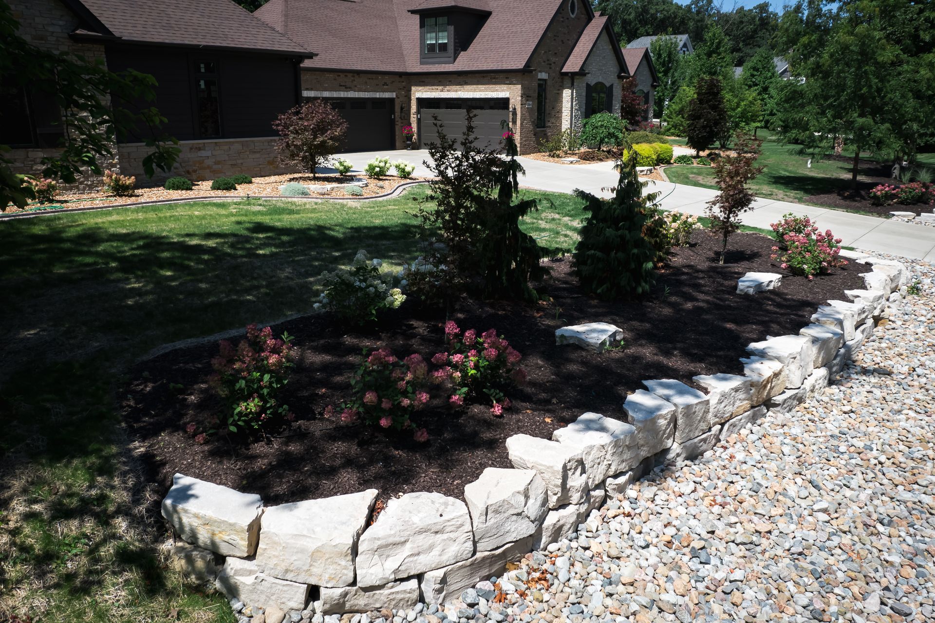 Front yard with stone-edged flower bed. House with brown roof and driveway.