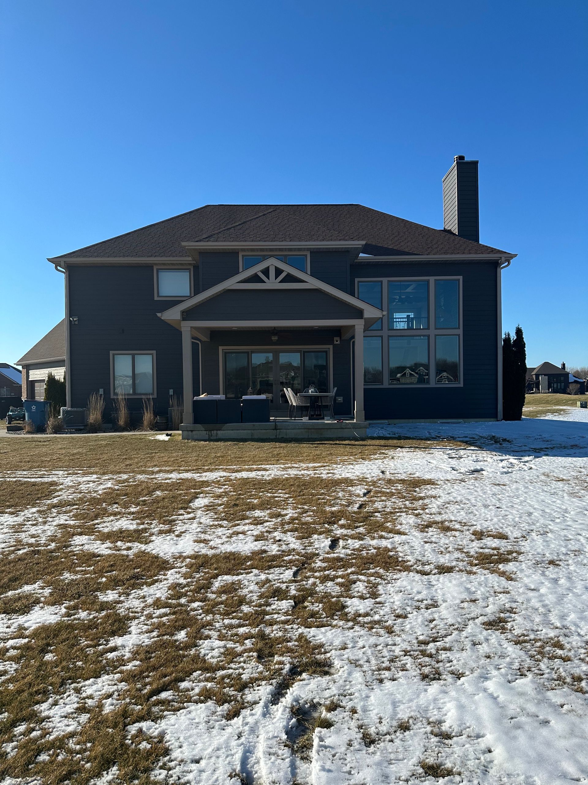 Back of a two-story blue house with a covered porch, windows, and a chimney on a snow-covered lawn under a blue sky.