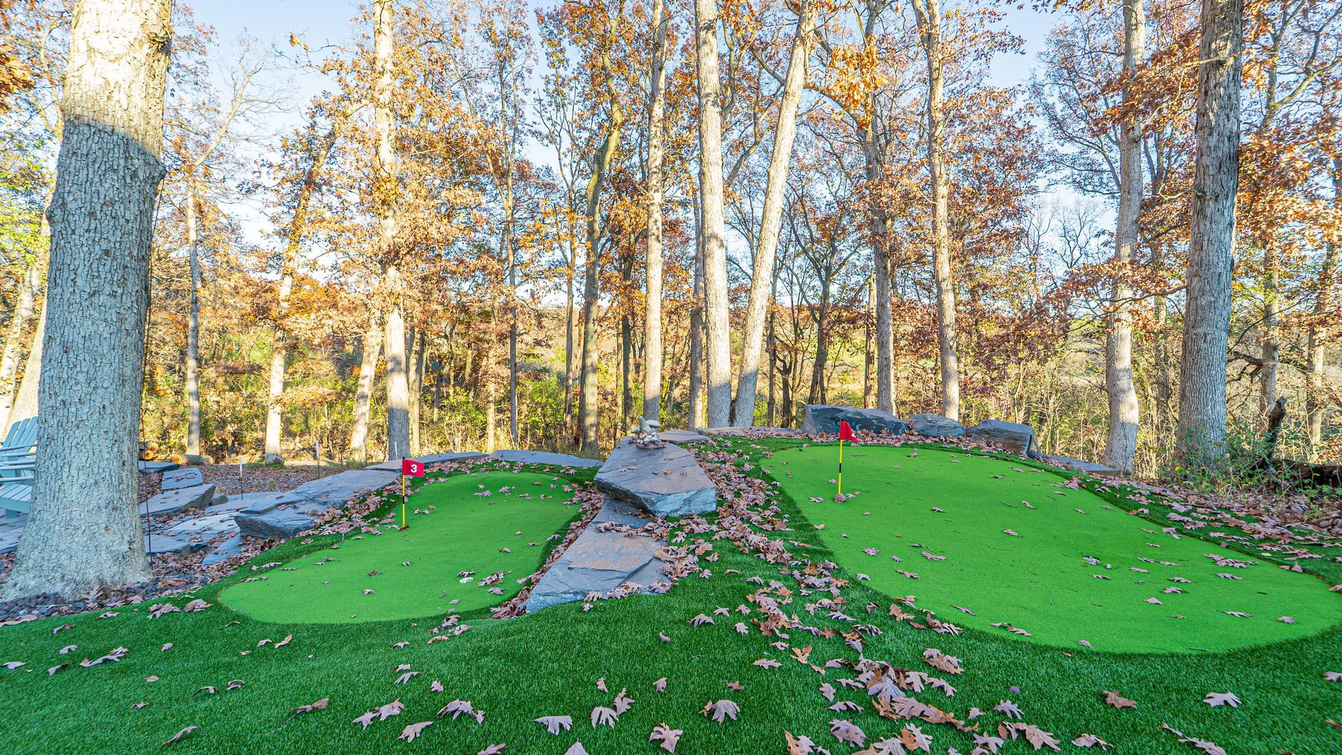 Two green artificial turf putting greens with red flags, surrounded by trees and rocks.
