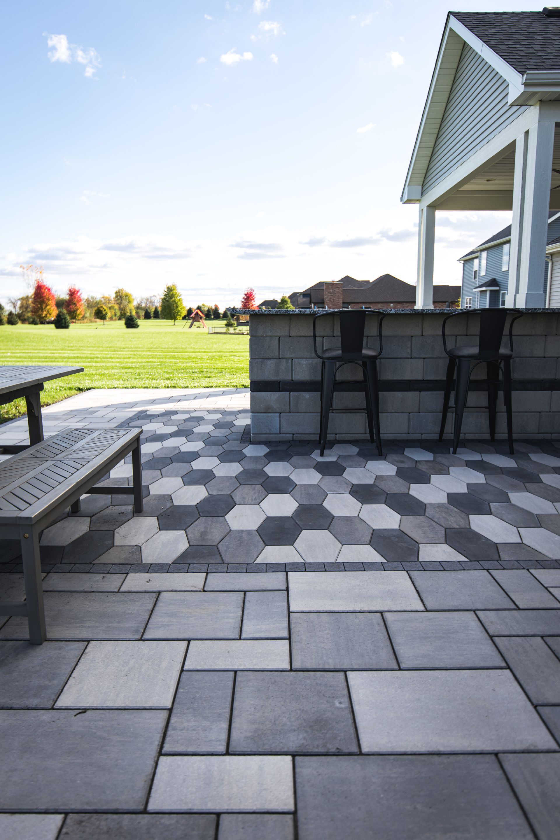 Patio with geometric pavers, outdoor bar, and chairs, with grassy lawn in background.