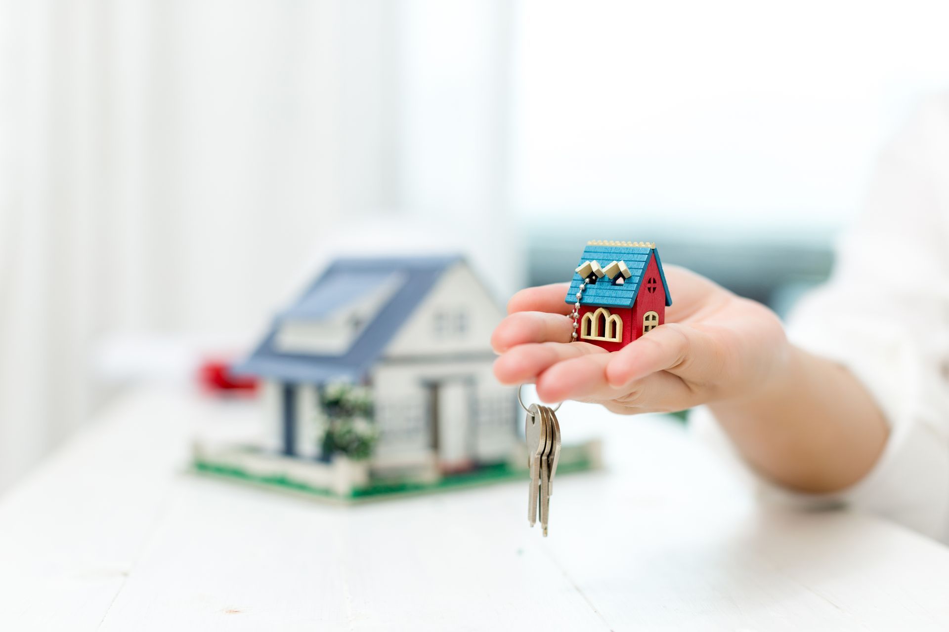 A hand holds a miniature house keychain above a blurred model house on a white surface.
