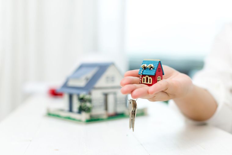 A hand holds a miniature house keychain above a blurred model house on a white surface.