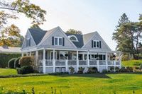 A light gray coastal-style house with dark shutters and a large wrap-around porch, set in a sunny, grassy yard.