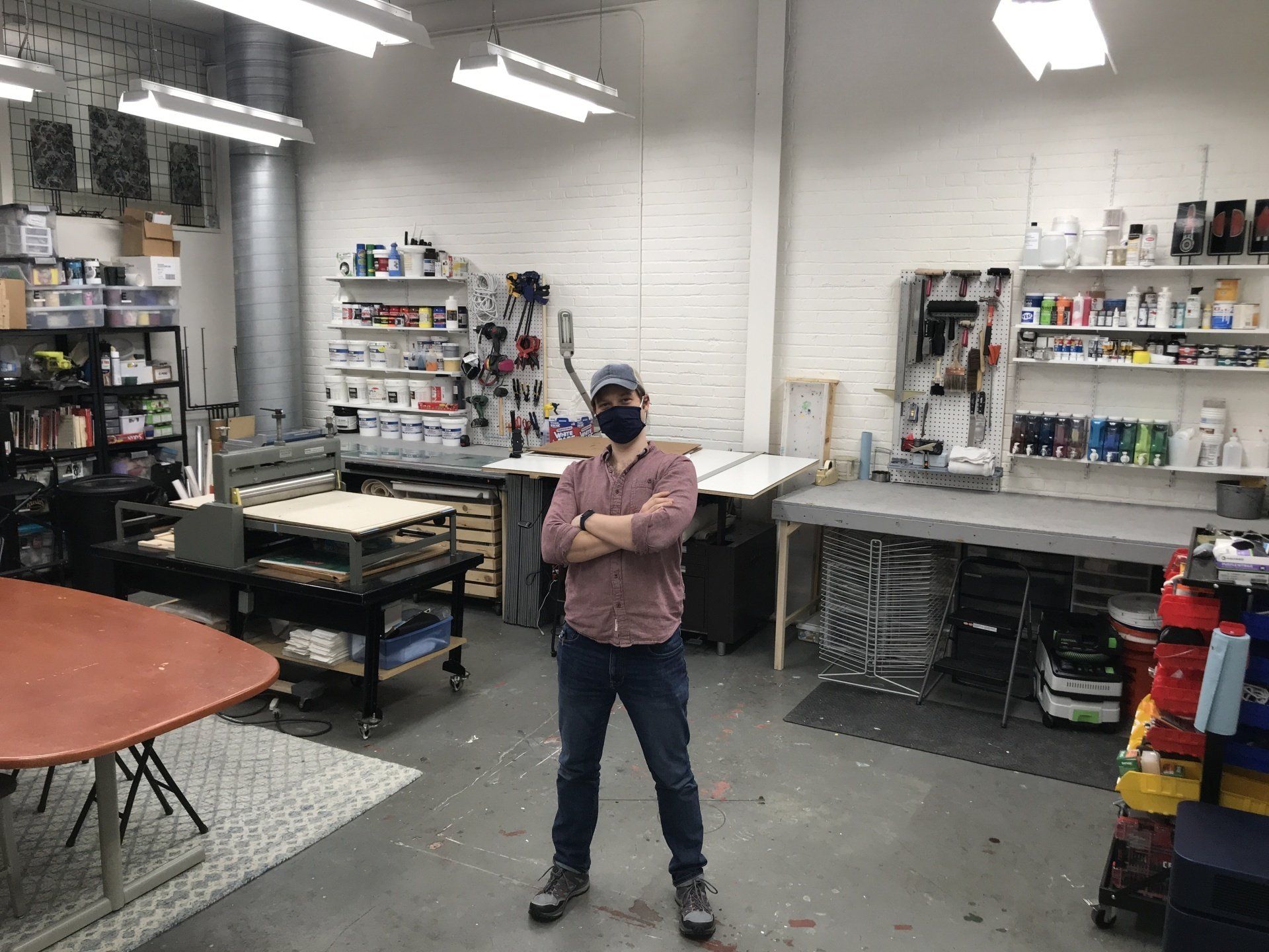 Garubba stands in proud posture at center of a wideshot of his studio, with tables, printing press, drafting table, tools, and paints.