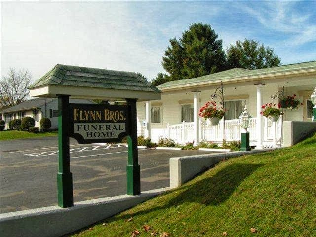 Funeral home building with green sign, white porch, and hanging flower baskets on a sunny day