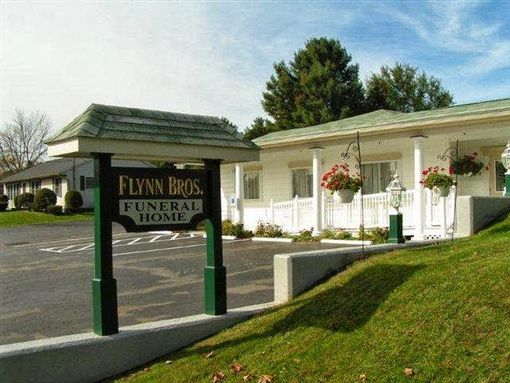 Funeral home building with green sign, white porch, and hanging flower baskets on a sunny day