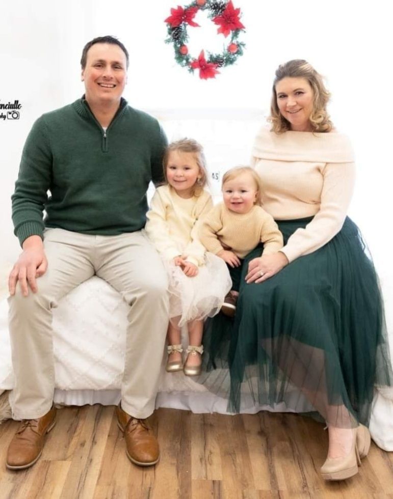 Family of four seated on a white couch, smiling in festive winter attire with a wreath on the wall.