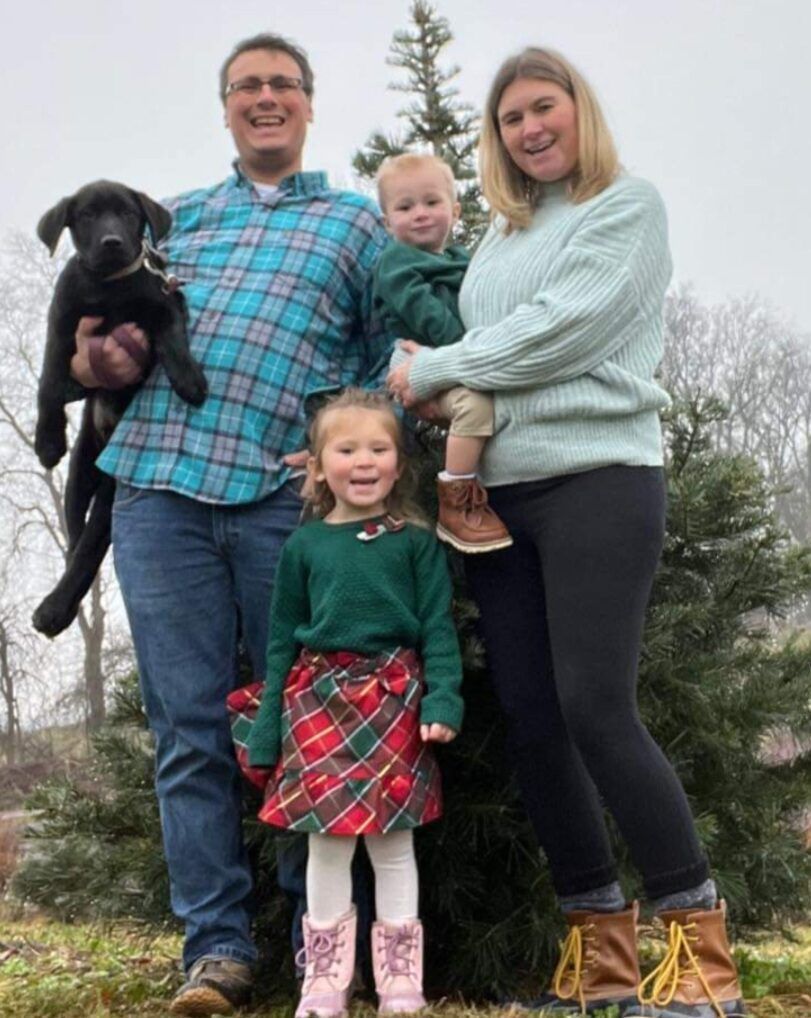 Family of four outdoors with a black dog, standing on grass in front of pine trees